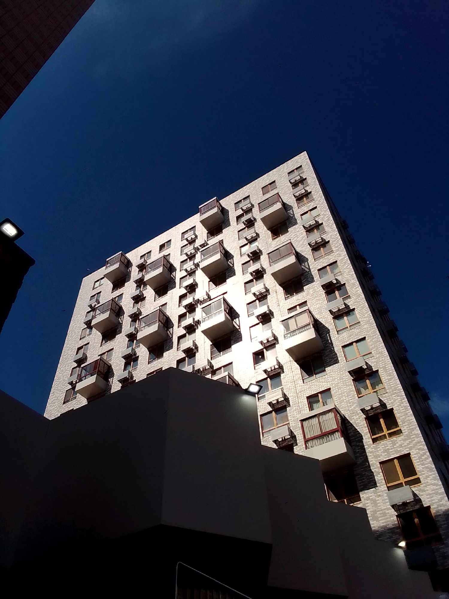 A low-angle, close-up shot captures a modern, white brick apartment building against a deep blue sky. The building has numerous balconies with dark railings and inset windows with wooden frames. A dark, angular structure is in the foreground, obscuring the lower portion of the apartment building and casting a shadow. A light fixture is visible on the left side of the dark structure. The composition emphasizes the geometric lines of the architecture and the stark contrast between the building and the sky.