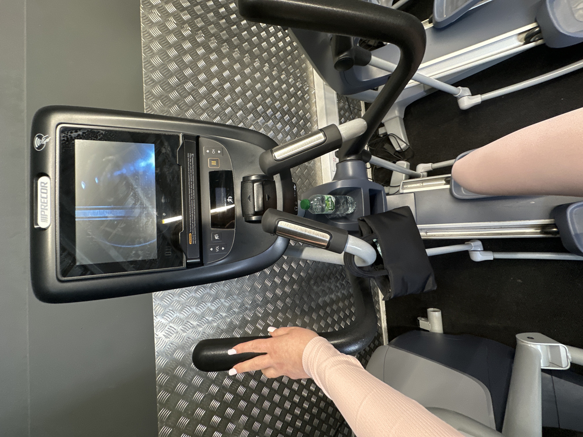 The image shows a close-up view of the console and handlebars of a Precor elliptical machine in a gym. A woman's hand with light pink nail polish is resting on the left handlebar. Her arm is covered by a light pink long-sleeved top. The console displays a blue-toned graphical interface, and a small bottle of water is visible in a holder below the console. The gym environment includes a textured metal wall in the background and black flooring. No specific location in Carlow, Ireland can be determined from the image.