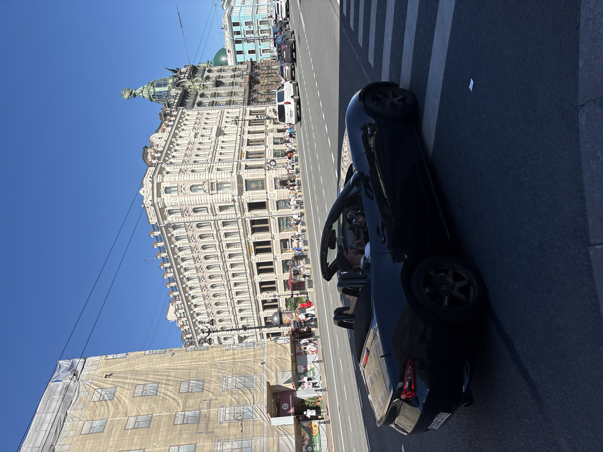 A black convertible Mazda Miata is parked at a pedestrian crossing on Nevsky Prospekt in Saint Petersburg, Russia. The car is facing right, and a man wearing a white shirt and a black baseball cap is visible in the driver's seat. In the background, several ornate buildings line the street, including the Singer House with its distinctive green dome and globe. People are walking on the sidewalk, and other vehicles are on the road. The sky is clear and blue. The building on the left is covered in scaffolding and a protective sheet.