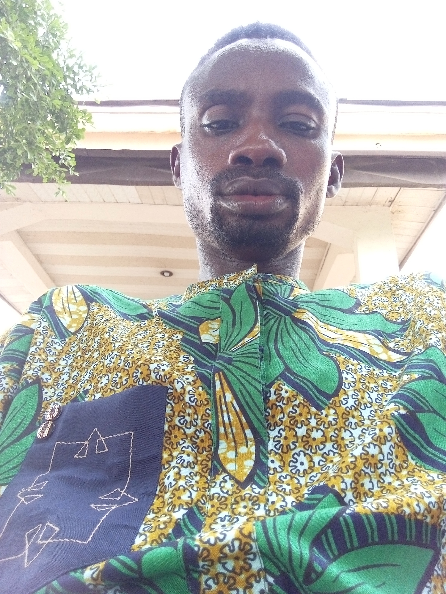 A selfie photograph shows an African man in an outdoor setting. The man has dark skin, a close-cropped haircut, and a trimmed beard. He is wearing a patterned shirt with green and gold floral and leaf designs. A dark blue patch pocket is visible on the shirt, adorned with light-colored stitching.
The man appears to be standing under a structure, possibly a covered porch or walkway, indicated by the presence of white beams or supports visible in the background. A tree with green foliage is partially visible in the upper left corner of the frame.
Based on the information provided, it's likely this image was taken in Lokoja, Nigeria, given that the user specified that this is the city where the photo was taken.