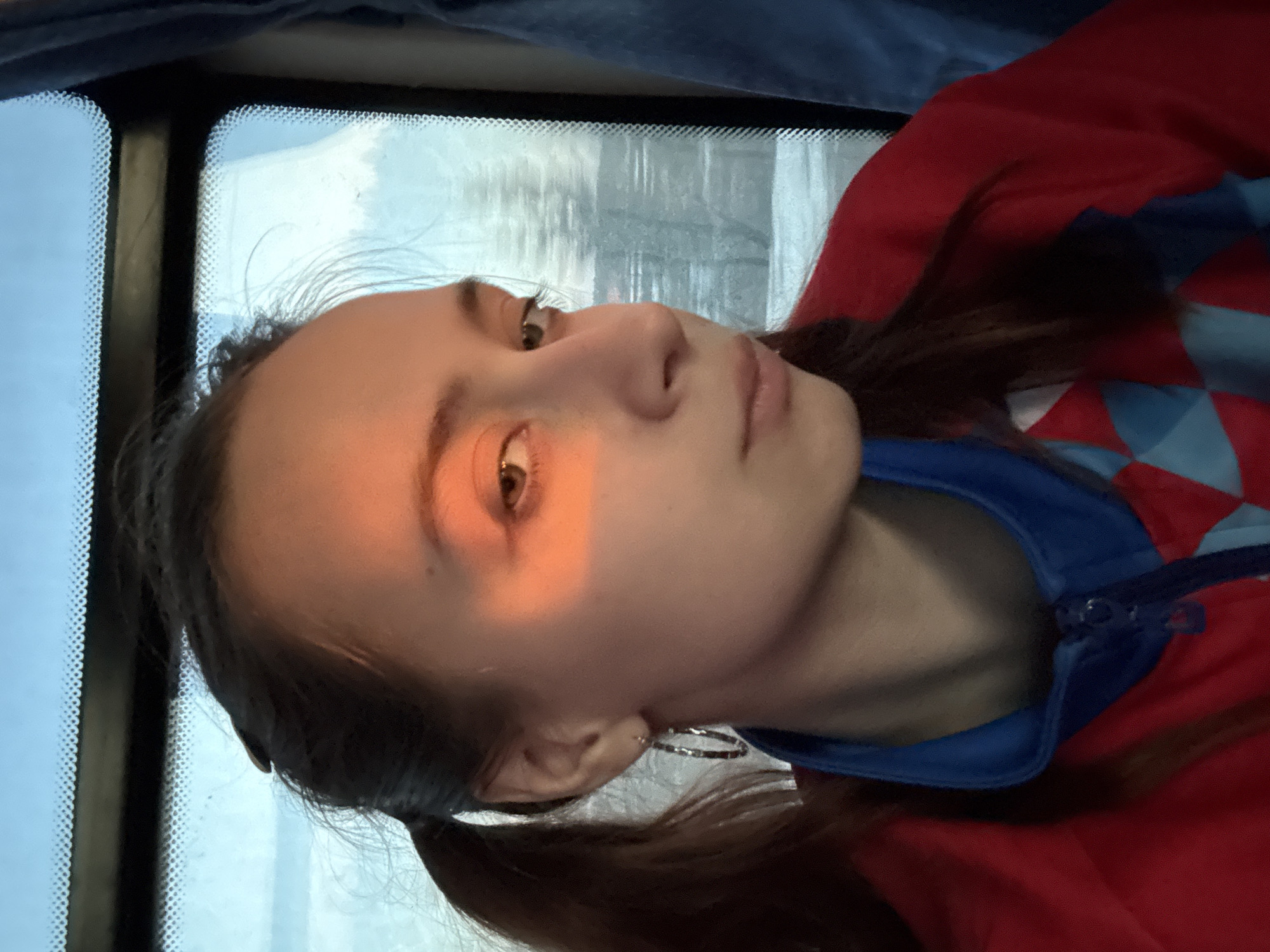 A young woman with light skin and brown hair in pigtails sits by a window with a blurry outdoor scene visible. She wears a red jacket with a blue collar. A bar of sunlight illuminates one side of her face. The image appears to be taken from a low angle, looking up at the subject. Location cannot be identified.