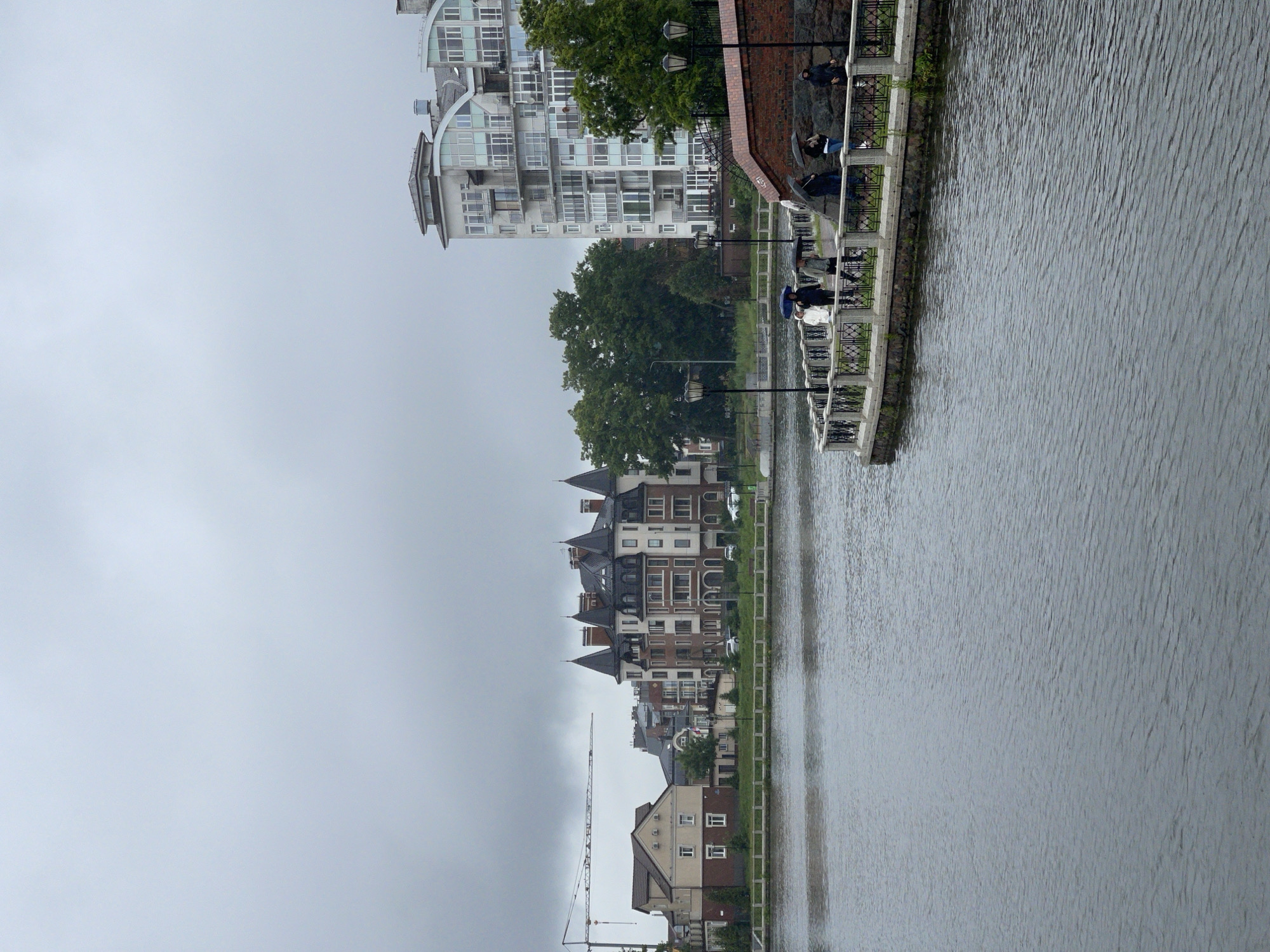 The image shows a waterfront scene in Kaliningrad, Russia, featuring a row of buildings along a canal. The buildings vary in architectural style, from modern apartment complexes to older, more traditional brick structures with steep roofs. Some buildings are tall and multi-story, with many windows, while others are lower and appear to be houses or smaller commercial buildings. A large construction crane is visible in the distance, suggesting ongoing development.
The canal is calm, with a textured surface that reflects the overcast sky. Along the edge of the canal, a walkway or promenade is visible, bordered by a decorative railing and lampposts. Several people are present on the walkway, some holding umbrellas, indicating it is raining or has recently rained. They appear to be strolling or gathered together. The weather is overcast and gloomy, contributing to a subdued atmosphere.
The overall scene captures a segment of urban life along the water, showcasing a mix of contemporary and historic architecture. The presence of people and the infrastructure of the walkway suggest a public space designed for leisure and enjoyment of the waterfront. The time of day is not explicitly indicated but the overcast sky suggests it is daytime. No visible text is present in the image.