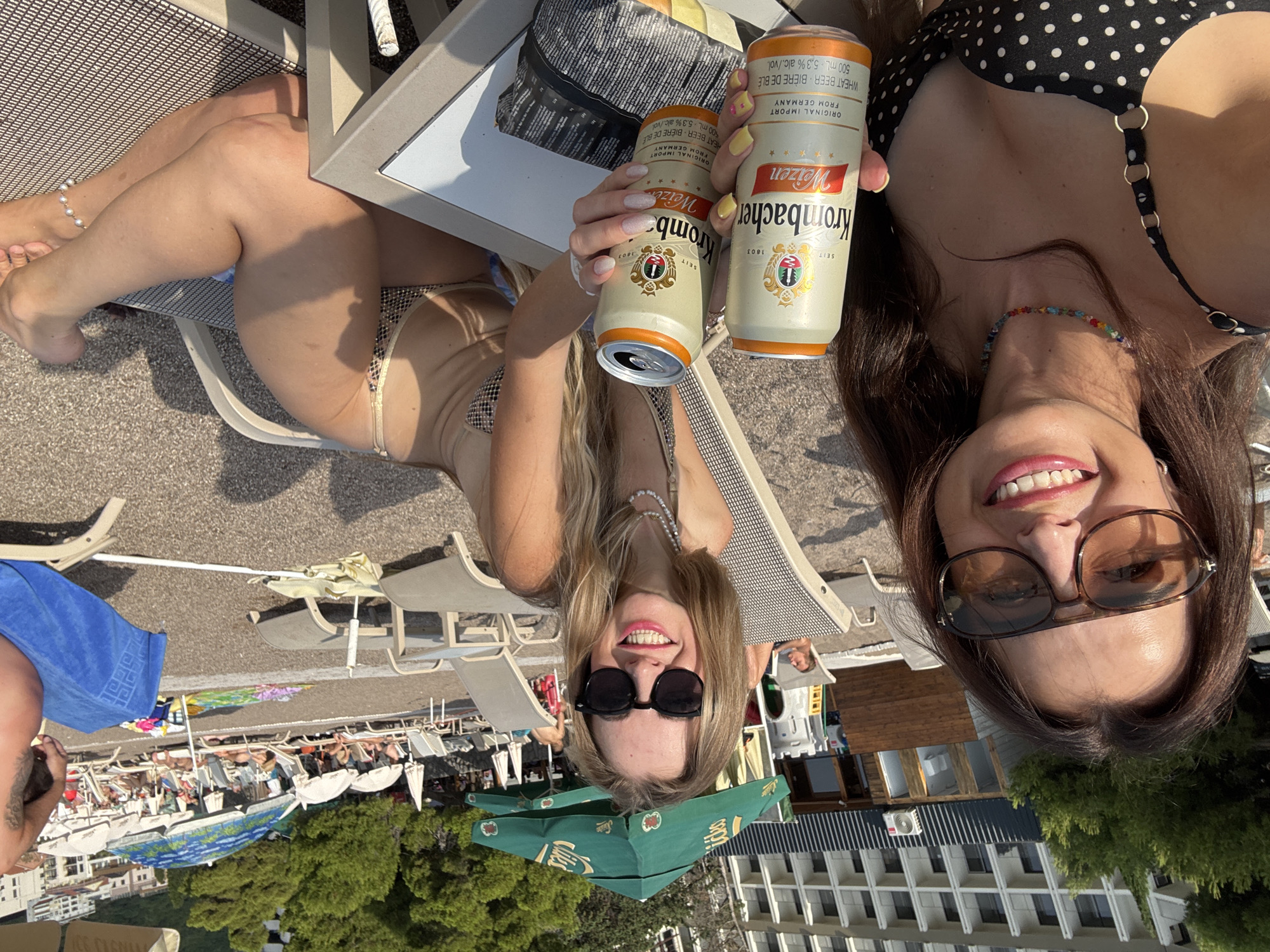 Two women are taking a selfie on a sunny day, likely at a beach resort. The woman on the left is lying down, wearing a bikini and an anklet. She is holding two cans of Krombacher Weizen wheat beer. The woman on the right, also wearing a bikini and sunglasses, is looking up at the camera with a wide smile. Her hair is dark brown, and she is wearing a colorful beaded necklace.
In the background, there are lounge chairs, beach umbrellas, and people relaxing. Buildings and trees are also visible, suggesting an outdoor recreational area. The overall mood is relaxed and festive, with the women appearing to enjoy their drinks and the sunny weather. The cans of Krombacher Weizen beer are clearly visible, with text indicating "ORIGINAL IMPORT FROM GERMANY" and "WHEAT BEER BIÈRE DE BLÉ 500 mL 5.3% alc./vol." The lighting suggests it is daytime, likely afternoon, given the strong shadows.