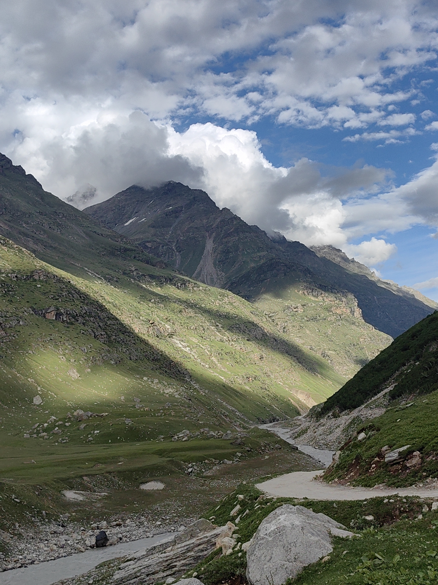 A sun-drenched mountain valley in Burwa, India, is captured in this vertical, eye-level shot. The scene is dominated by the majestic, green, and rocky slopes of the Himalayas, with sharp, dark shadows cast by clouds that are breaking up to reveal a bright blue sky. A winding river flows through the valley floor, its water a muted gray. The terrain is a mix of lush green grass, scattered rocks, and exposed earth, with a dirt road visible on the right side of the frame. The weather appears to be a mix of sun and clouds, suggesting a pleasant day in the mountains. No people or animals are visible in the image.