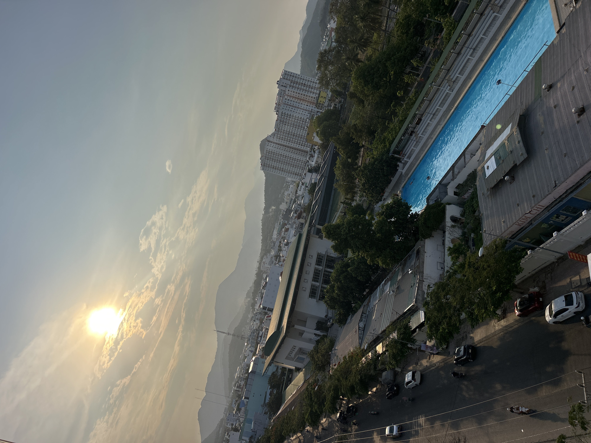 The image shows an elevated view of a section of Bac Nha Trang Ward, Vietnam. The sky is light with a bright sun obscured by haze. Mountains are visible in the distance. Closer to the camera, tall apartment buildings stand. A long, narrow blue pool is located on top of a building. A street with cars and motorcycles is visible along the bottom. Buildings and trees populate the foreground and middle ground.