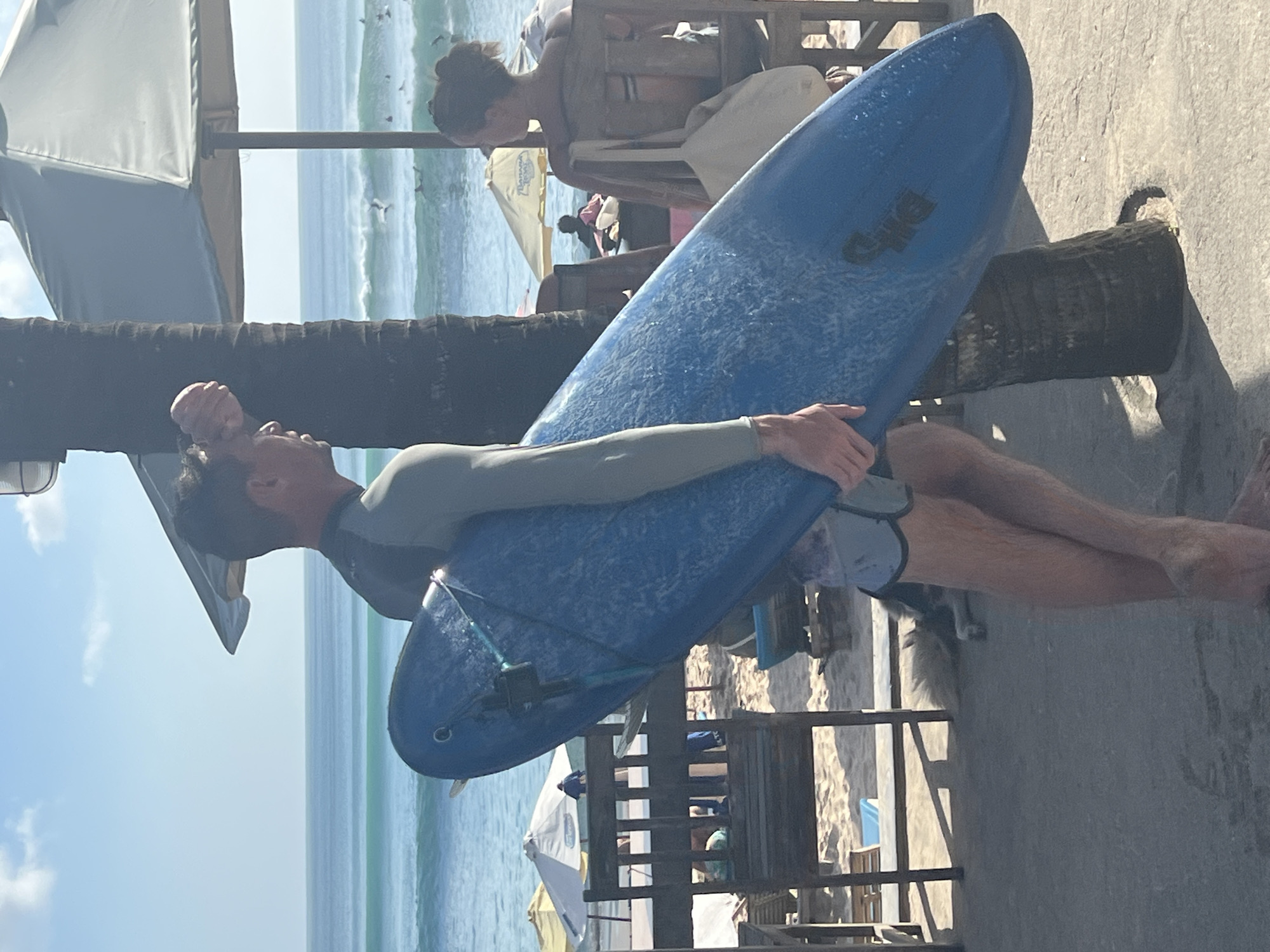 On a sunny day in Kuta Selatan, Indonesia, a man is carrying a blue surfboard. He is wearing a long-sleeved grey rashguard and patterned board shorts. The surfboard is resting on his shoulder and held in place by his arm. In the background, the ocean meets the beach, where people sit under umbrellas. A couple of people are seated on chairs. A palm tree trunk is behind the surfboard.