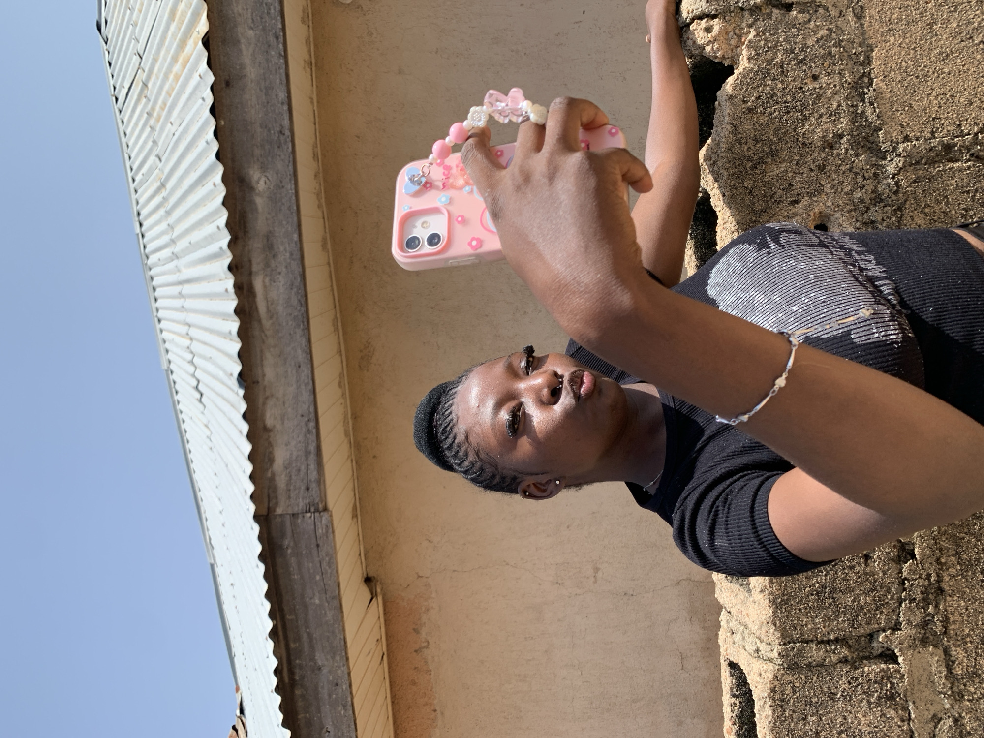 A young Black woman with braided hair pulled into a bun is taking a selfie outdoors in Jos, Nigeria. She is wearing a black ribbed top and a silver bracelet on her left wrist. Her phone, in a pink case adorned with charms, is held in her right hand. The background consists of a textured tan wall, a brick structure, a corrugated metal roof, and a clear blue sky. She has her lips pursed as if for a kiss.