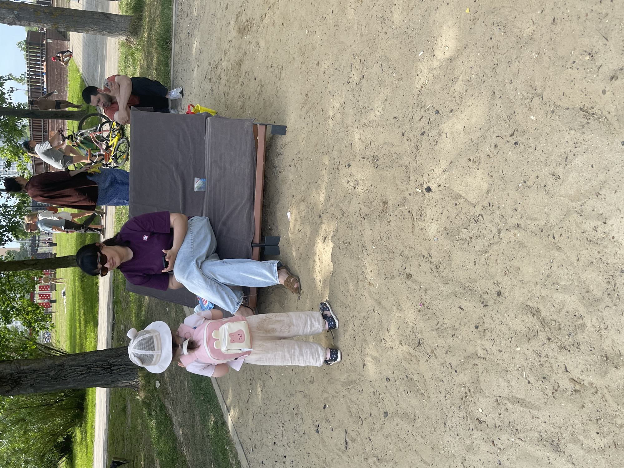 This image depicts an outdoor scene within a public park or recreational area in Moscow, Russia, captured during daylight.
In the immediate foreground, a wide expanse of light-colored sand, marked with small debris, occupies the lower half of the frame. A dark gray upholstered sunbed with a brown base is positioned horizontally in the mid-left. On the sunbed, a small clear package with colored contents is visible.
A young child, with their back to the camera, stands on the sand, facing the sunbed. The child wears a light-colored mesh bucket hat with ear-like protrusions, a light pink t-shirt, light beige long pants (with some visible dark smudges), and dark sandals adorned with white patterns. A light pink backpack featuring a white bunny face and ears is worn on the child's back.
Seated on the sunbed, partially visible, is an adult female. She has dark hair, wears dark sunglasses, a short-sleeved purple t-shirt, light blue jeans, and brown open-toed sandals. She is looking towards the child and holding a smartphone in both hands.
To the right of the sunbed, an adult male crouches on the sand, facing towards the left. He wears a reddish-brown t-shirt, dark pants, and light-colored sneakers, and is holding a smartphone in his right hand. A yellow bucket with a red handle is partially visible near the sunbed's right end.
The background features a vibrant green park environment with numerous mature trees casting shadows. Paved pathways meander through grassy areas. Several other individuals are visible in various activities:
* An adult female stands near the left side of the sunbed, facing away, wearing a brown top and blue jeans.
* A person rides a bicycle further back.
* Another person stands without a shirt, wearing dark swim trunks, near a wooden fence.
* Other park-goers are walking or standing in the distance.
* In the far background, apartment buildings and playground structures with red and white elements are visible.