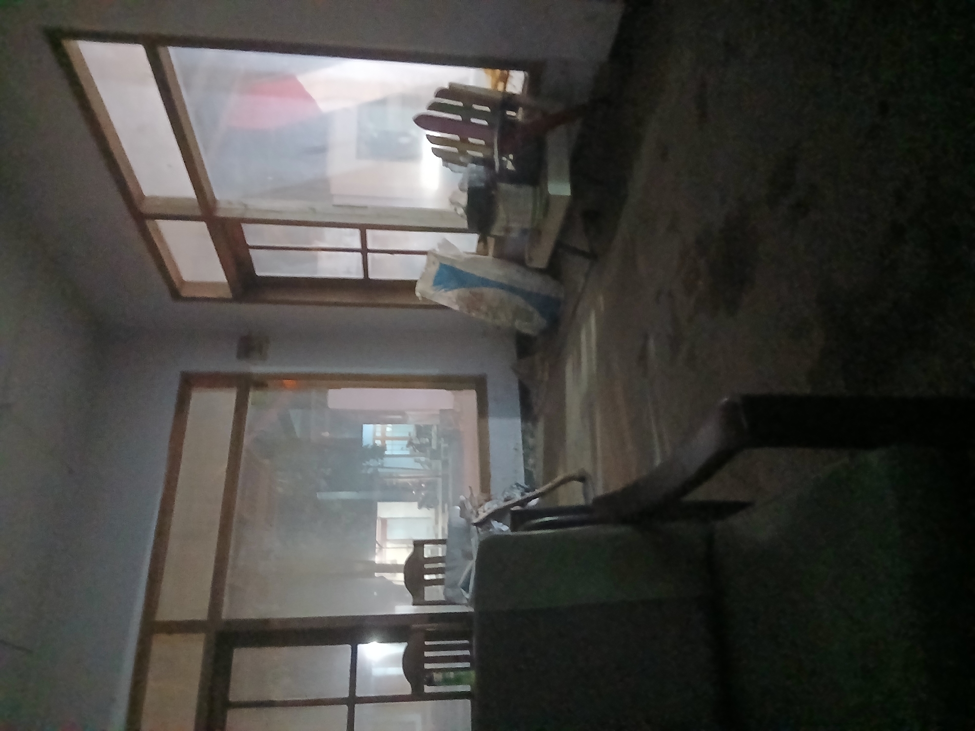 The image depicts a dimly lit interior space with large windows offering a view of an outdoor area, possibly Kademangan Kulon, Indonesia. The room appears to be an unfinished or disused part of a building. In the foreground, a portion of a dark-colored chair or sofa is visible on the left. The floor is covered with dirt or debris, and there are several objects scattered around.
To the right of the foreground seating, there are two wooden chairs, one with a slatted back. A large, white and blue bag, possibly containing building materials, leans against the wall near a table. On the table, there is a bucket and other unidentifiable items, along with a second wooden chair.
Through the windows, the outdoor scene shows more building structures and what appear to be motorcycles or scooters parked outside. The lighting suggests it is either dusk or nighttime, with artificial lights visible in the outdoor area. The overall impression is one of a neglected or under-construction space.