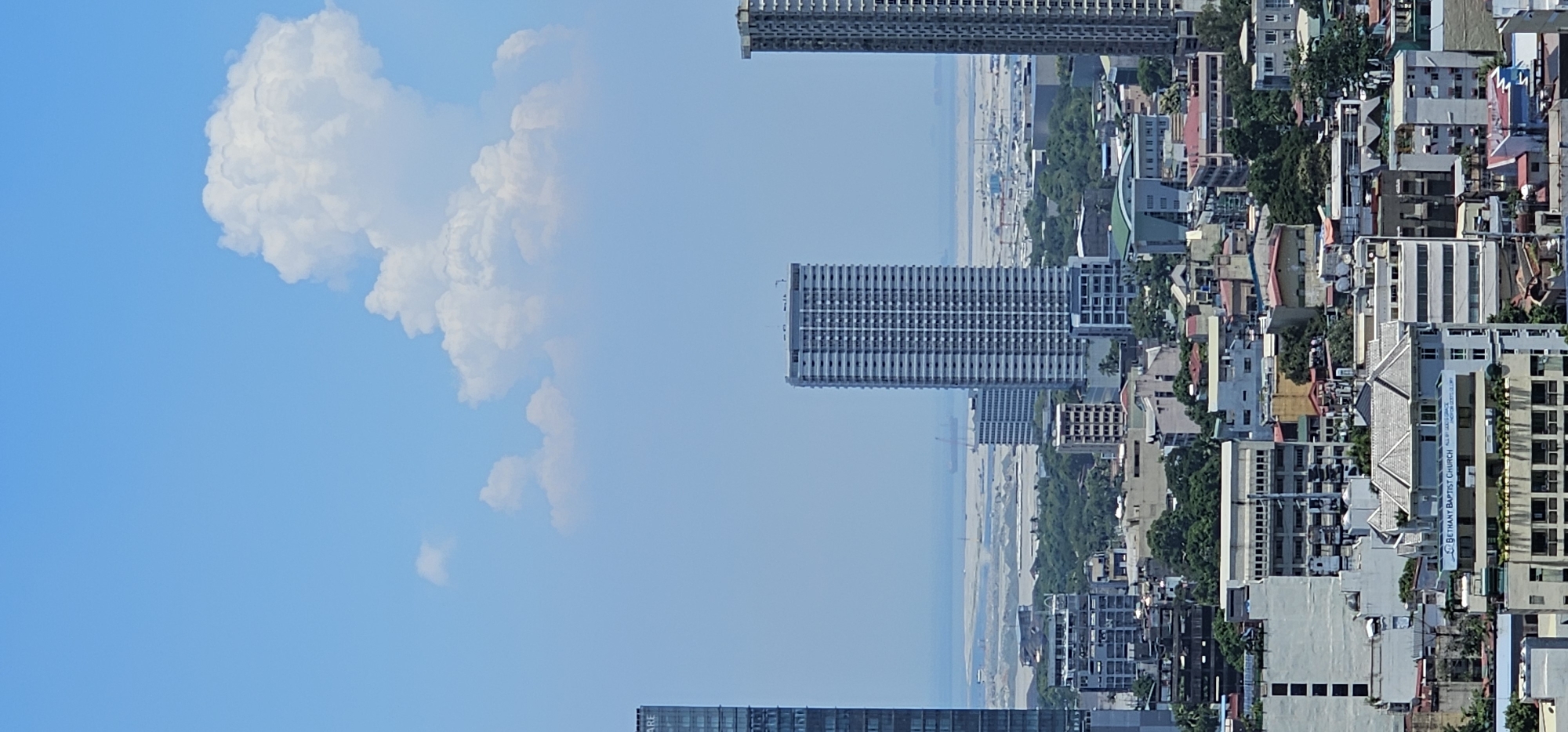 This is a telephoto, eye-level, vertical, outdoor, daytime, full shot of a sprawling cityscape, featuring a dense collection of buildings of various sizes and architectural styles. The image is dominated by the clear blue sky, with a large, fluffy white cloud positioned towards the top right quadrant. In the middle ground, several tall, modern high-rise buildings stand out, characterized by their repetitive window patterns and smooth facades. Further back, the urban sprawl continues, with smaller buildings and clusters of trees creating a textured landscape. In the distant background, beyond the city, a hazy horizon suggests the presence of water or a flat, open area, with a ship or large vessel visible on the water, possibly indicating a port or industrial zone. A prominent building in the lower center of the image bears a sign that reads "BETHANY BAPTIST CHURCH." The scene is devoid of any visible people or distinct actions, presenting a static, architectural overview of a developed urban environment. The overall impression is one of a densely populated, modern metropolitan area.