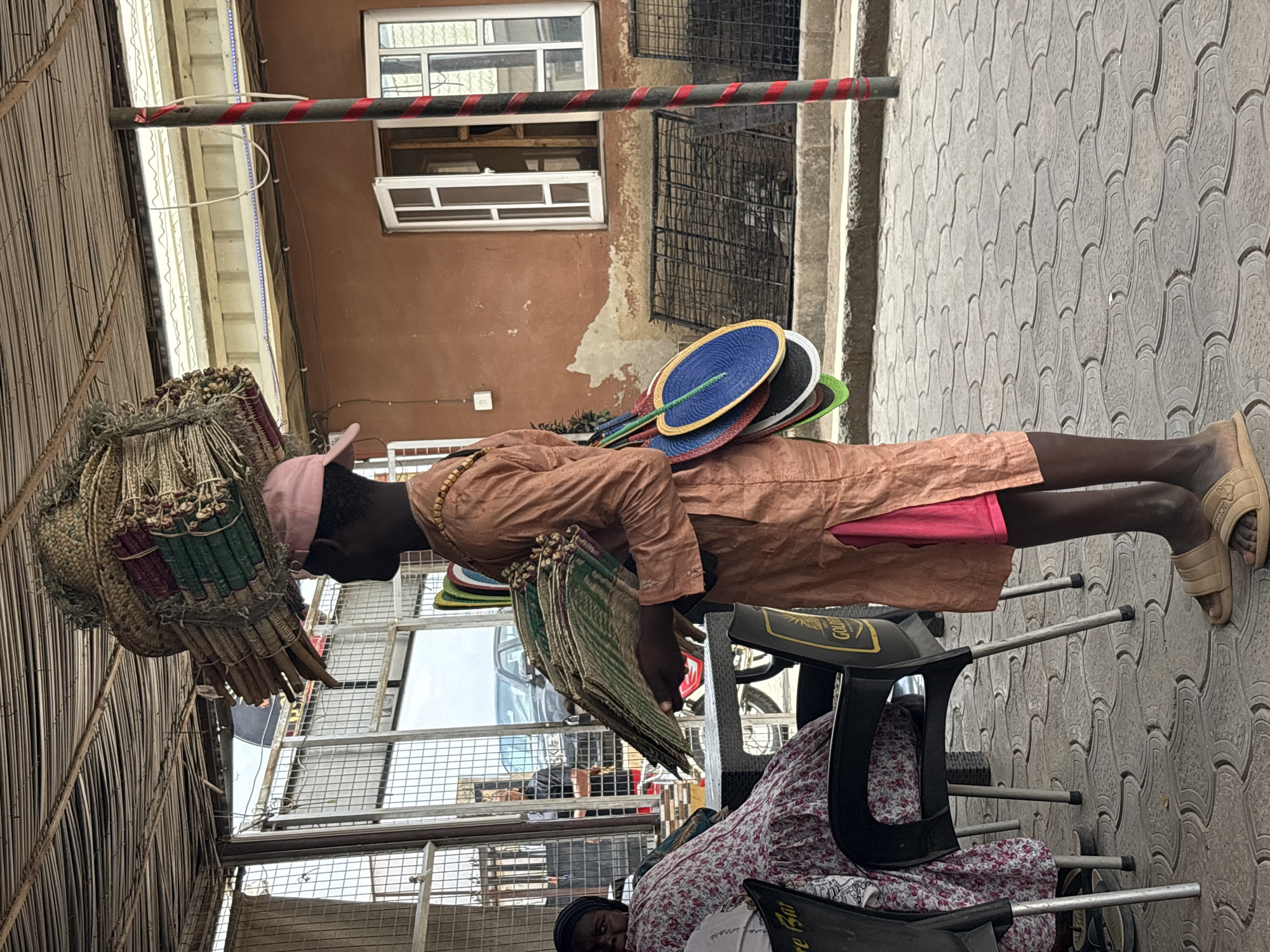 The image depicts an outdoor street scene, likely a bustling market area, in Abuja, Nigeria. The primary focus is a dark-skinned individual standing upright, acting as a vendor. They wear a light brown or tan long-sleeved tunic over what appears to be a pink garment and a pink cap.
The vendor is laden with goods for sale. On their head, they balance a large, intricately woven straw-like structure, possibly a type of headpiece or basket, which is filled with numerous smaller, tightly rolled items, perhaps mats or fans, secured with colored wraps (green, red, light brown). Tucked into their back are several colorful, circular hand fans in shades of blue, black, white, and green. In one arm, they carry a stack of flatter, rectangular woven mats or fans. The vendor wears light-colored, open-toed sandals.
In the lower right foreground, partially obscured, another person is seated on a black plastic chair with metal legs. This person wears a floral-patterned dress or top and has dark hair or a head covering. The back of another black plastic chair is visible, featuring a clear "Gulder" logo, a well-known Nigerian beer brand, further confirming the location context.
The ground is paved with interlocking grey pavers in a patterned design. In the background, there is a building with a reddish-brown facade. It features white-framed windows, some with metal security grilles. An overhead structure, possibly a thatched or woven bamboo roof, provides shade on the left side of the frame, with long, bright white and blue LED light strips attached beneath it. A horizontal metal bar wrapped with red and white striped tape (resembling caution tape) is also visible, extending across the upper part of the scene. The overall impression is a daytime setting, focused on street commerce.