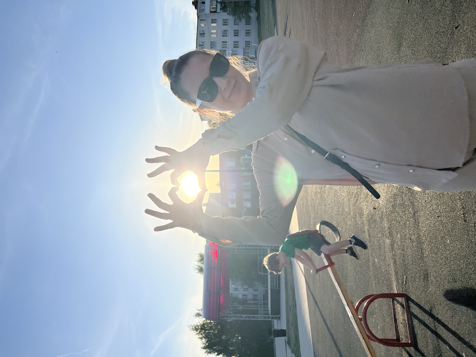 The image shows a woman making a heart shape with her hands around the sun. She wears sunglasses and a light-colored shirt. A child is upside down on a seesaw. A stage structure is visible in the background, along with a residential building. The location is Noviy, Russia. The image is taken during a sunny day.