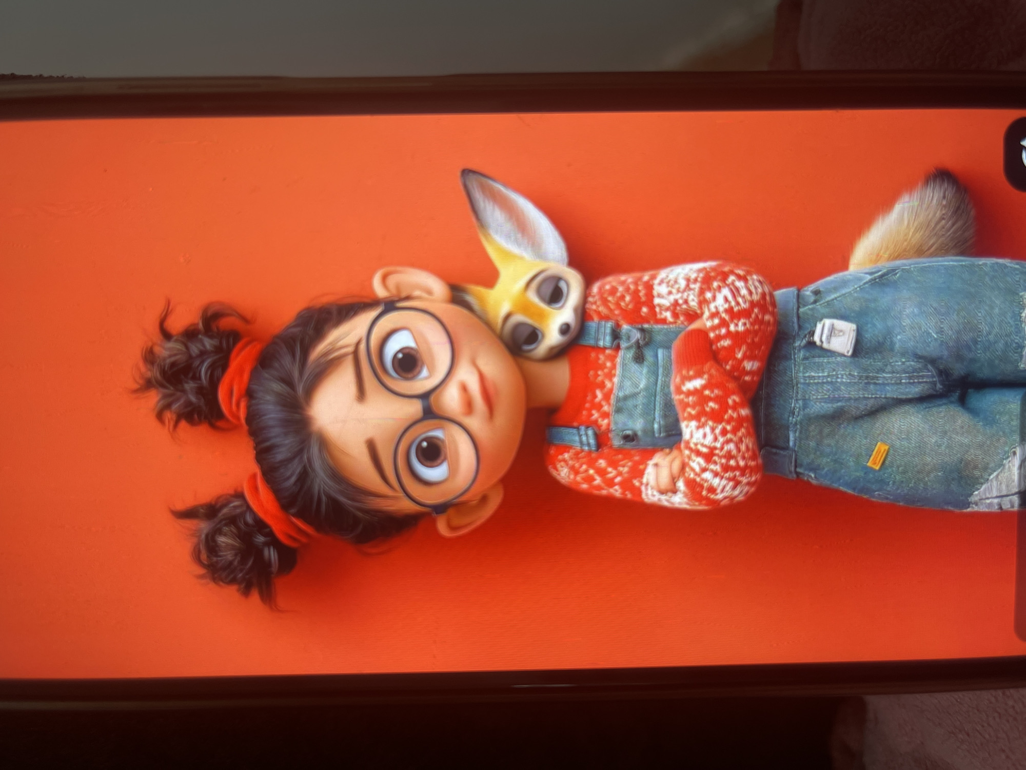 A digital illustration of an animated young girl with dark hair styled in two messy buns, wearing red scrunchies, large round glasses, and a red and white patterned sweater with denim overalls. She has her arms crossed and a small fennec fox rests on her left shoulder. The background is a solid, vibrant orange. The image is displayed on a phone screen, with a portion of the phone's bezel visible on the left and right edges. There is no indication of the city of Bishkek City, Kyrgyzstan, or any real-world environment.