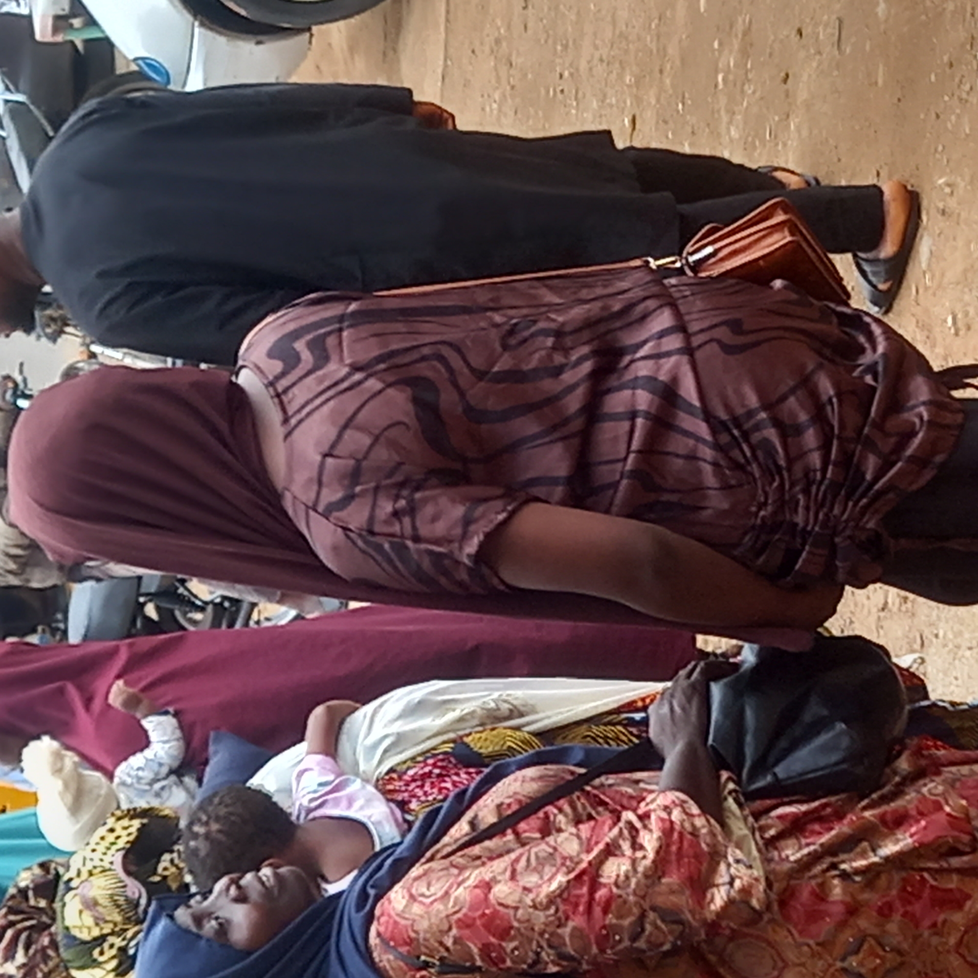 The image shows several people, possibly vendors, in a market or public area in Dadi Riba, Nigeria. One woman is lying down, partially covered in fabric with a child nearby. Other individuals are standing near or passing by. A person in a dark coat and shoes, carrying a brown bag, is visible in the background. A motorcycle or scooter is also present. The ground is dusty or unpaved.