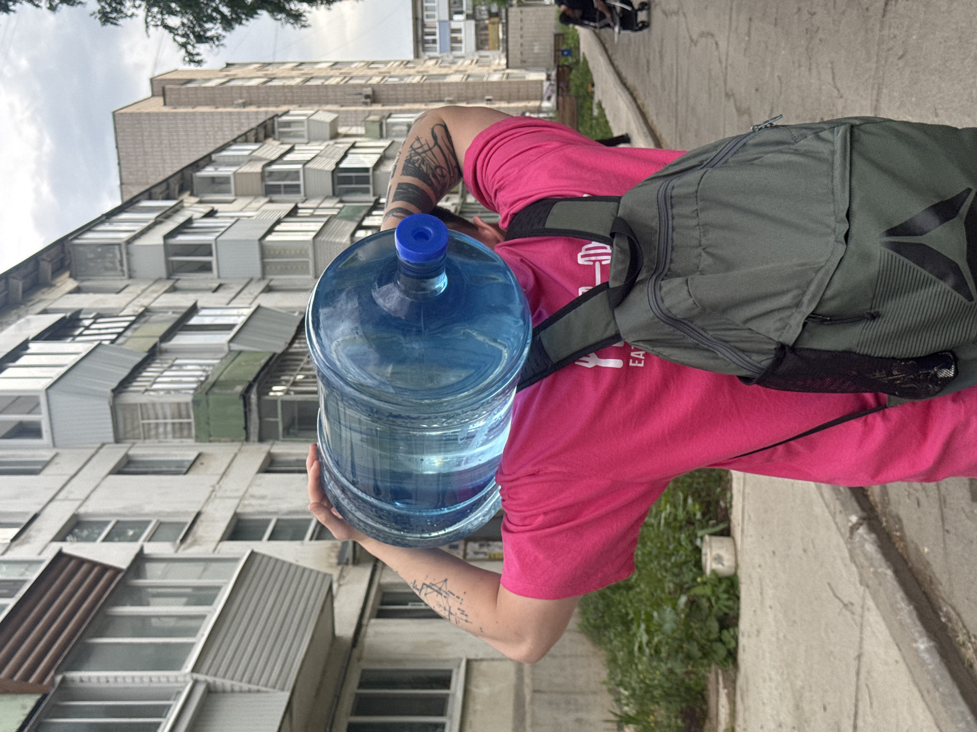 A person in a pink shirt is carrying a large, blue water bottle on a city street in Chaikovsky, Russia. The person is also wearing a green backpack. The background shows a tall, older apartment building with numerous windows and metallic sunshades. A sidewalk runs alongside the building with a curb separating it from a grassy area. Another person can be seen in the background.