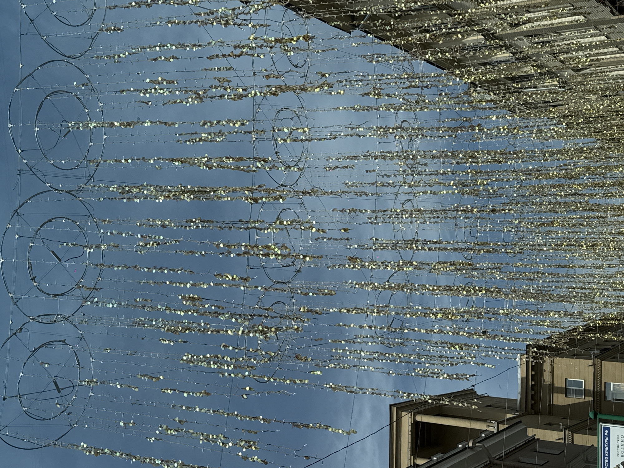 The image, captured in Moscow, Russia, presents an upward view of an urban street or public space adorned with numerous decorative elements suspended overhead against a bright blue and lightly clouded sky. The main subjects are countless horizontal strings of light-colored, possibly reflective or translucent, leaf-shaped decorations, resembling garlands or fairy lights. These strands stretch across the entire frame, creating a shimmering canopy effect.
Interspersed with and supporting these garlands are several large, open-frame circular metallic structures, primarily visible on the left side of the image. These circles, arranged in vertical series, suggest part of a complex overhead lighting or display system.
In the background, on the right and bottom edges, parts of multi-story urban buildings are visible, characterized by their windows and architectural details, confirming the city setting. The natural daylight indicates it is daytime, and the decorative lights appear to be off or are not fully illuminated, blending with the bright conditions. The clear sky suggests fair weather.
A notable detail is a partially visible green and blue sign on a building in the bottom right corner. Cyrillic text on the sign includes "РСТВО КУЛЬТУРЫ РФ" (part of "Ministry of Culture of the Russian Federation"), "РОССИЙСКОЕ" (Russian), and "ВОЕННО" (Military), indicating a governmental or cultural institution. The overall impression is one of festive urban decoration, possibly for a holiday or special event.