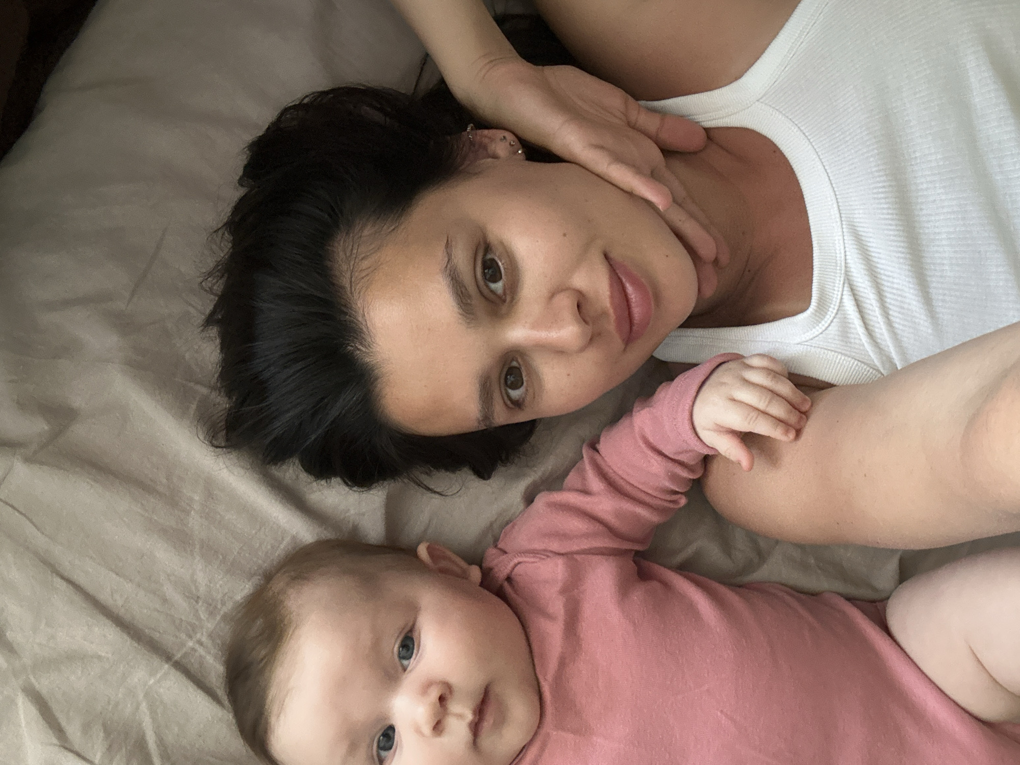 A high-angle, close-up photograph depicts a woman and a baby lying on what appears to be a bed. The woman has dark hair and is wearing a white tank top. Her hand is touching her face. The baby, wearing a pink long-sleeved garment, lies next to the woman, looking towards the camera. The bed is covered with beige fabric.
There is no indication of the location being in the city of Chaikovsky, Russia, within the image.