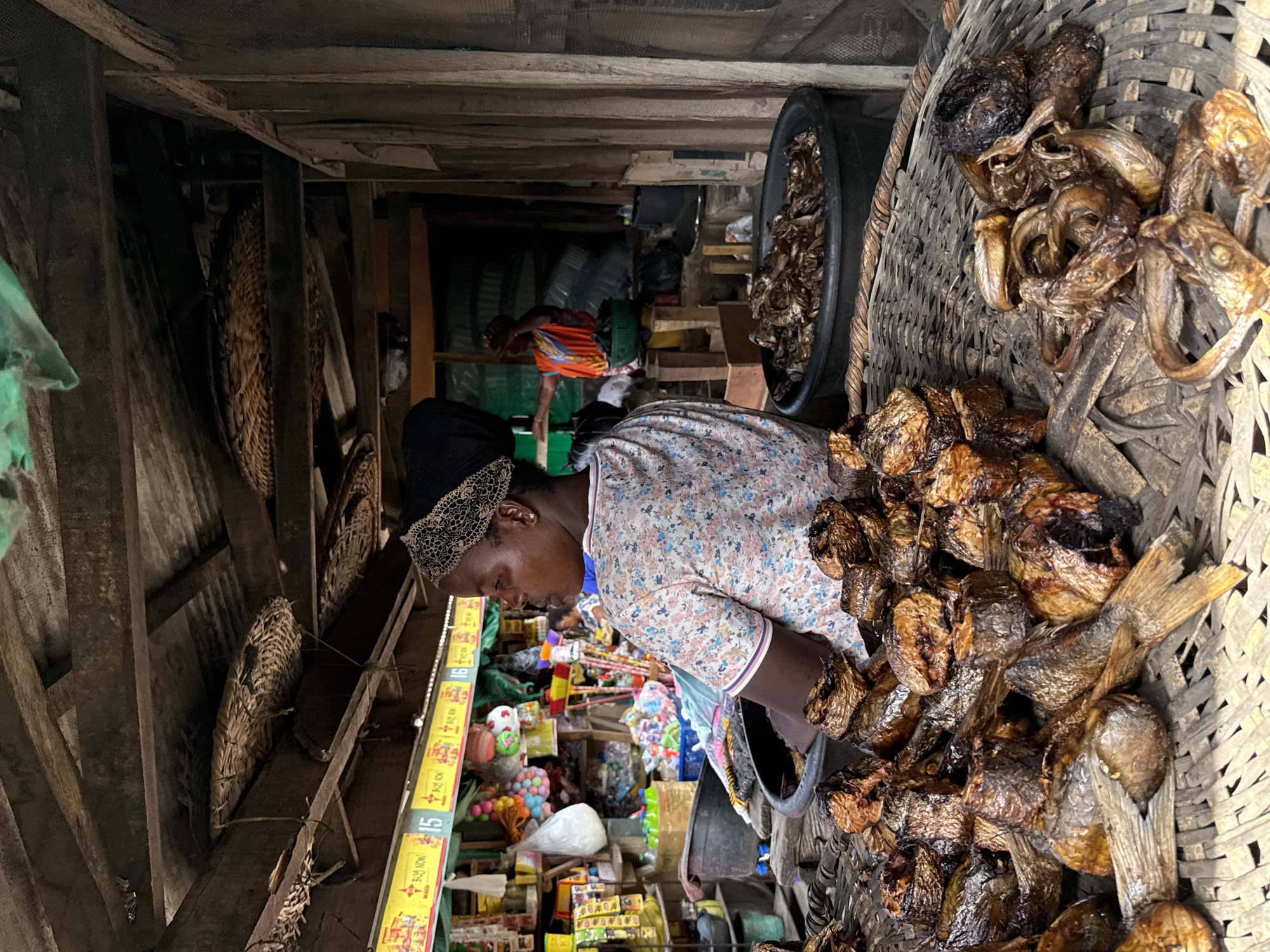 A woman wearing a floral top and head covering tends to a large basket of smoked fish in an indoor market. She leans toward the basket, possibly arranging or selecting the fish. A black tub with more smoked fish sits beside the basket. The market appears crowded, with various goods on display, including toys and packaged food items. In the background, another person wearing an orange top is visible. Baskets are mounted on the ceiling. The image is likely taken in a market setting in Lekki, Nigeria.