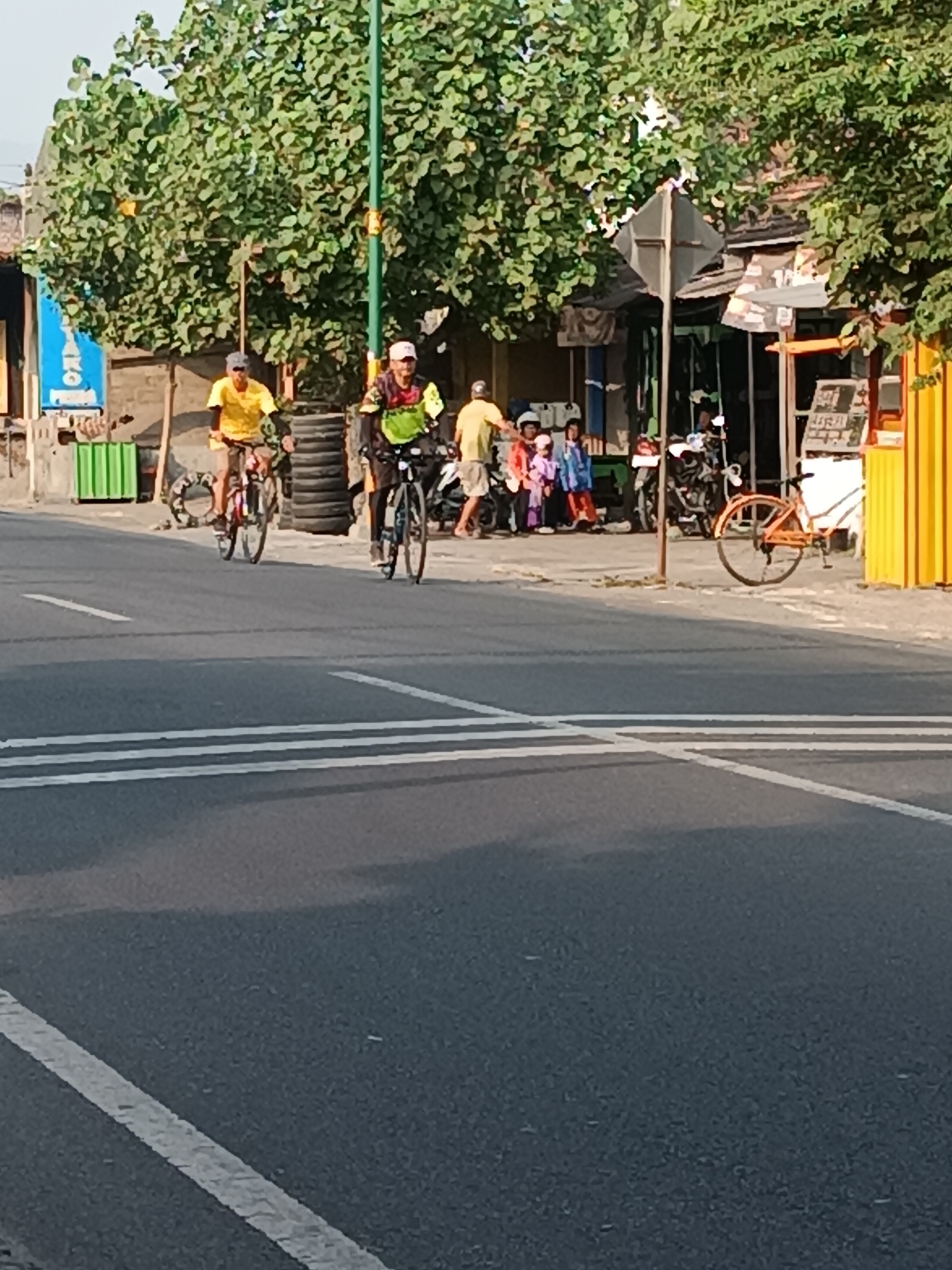 In Imogiri, Indonesia, this image captures a street scene with several individuals engaged in various activities. Two men are cycling on the asphalt road, one wearing a yellow shirt and the other a colorful cycling jersey. In the background, a group of people, including children, are gathered near shops lining the street. The environment appears to be a mix of commercial and residential buildings, with a lush green tree providing shade. Several parked motorcycles suggest a common mode of transportation in the area. An orange bicycle is leaning against a pole near the curb. The road has visible white lane markings. The lighting indicates it is daytime, possibly late morning or afternoon.