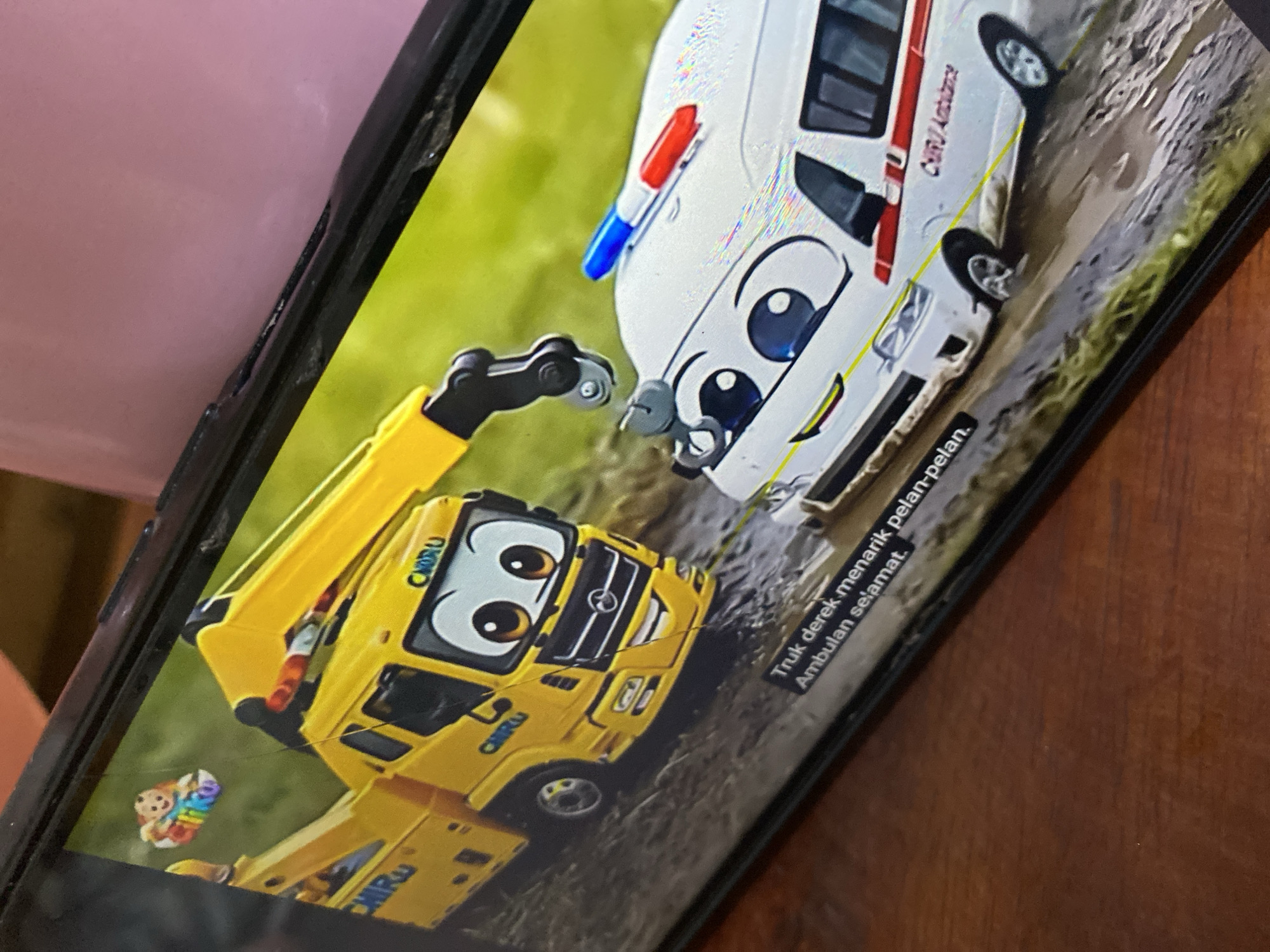 A yellow tow truck with a smiling face is helping a white ambulance, also with a smiling face, out of a muddy area. The tow truck's crane is hooked onto the ambulance. There are some green grassy areas in the background. The scene appears to be from an animated children's show. Text in Malay, "Truk derek menarik pelan-pelan. Ambulan selamat." which translates to "The tow truck pulls slowly. The ambulance is safe." is visible at the bottom of the screen. The image is a close-up of a smartphone screen displaying this animation.