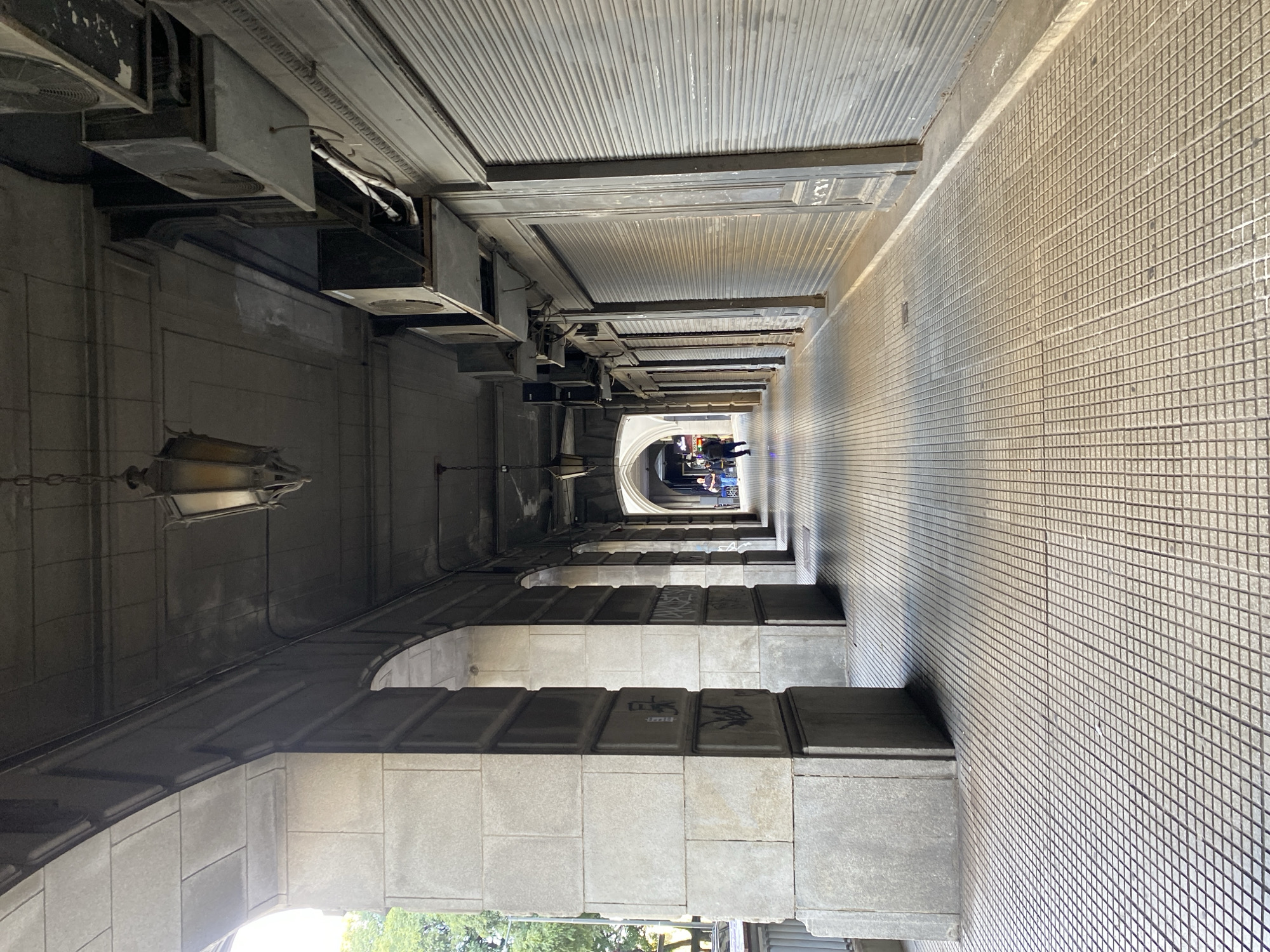 A long, narrow, outdoor corridor with a barrel-vaulted ceiling leads into the distance. The corridor is defined by gray stone arches on the left and a tiled wall on the right. A single hanging lamp is visible on the left. Mechanical equipment is affixed to the ceiling on the left side of the corridor. Two figures stand at the far end of the corridor. Green foliage is visible outside the opening in the foreground. The scene is set in a colonnade in the city of Buenos Aires, Argentina.