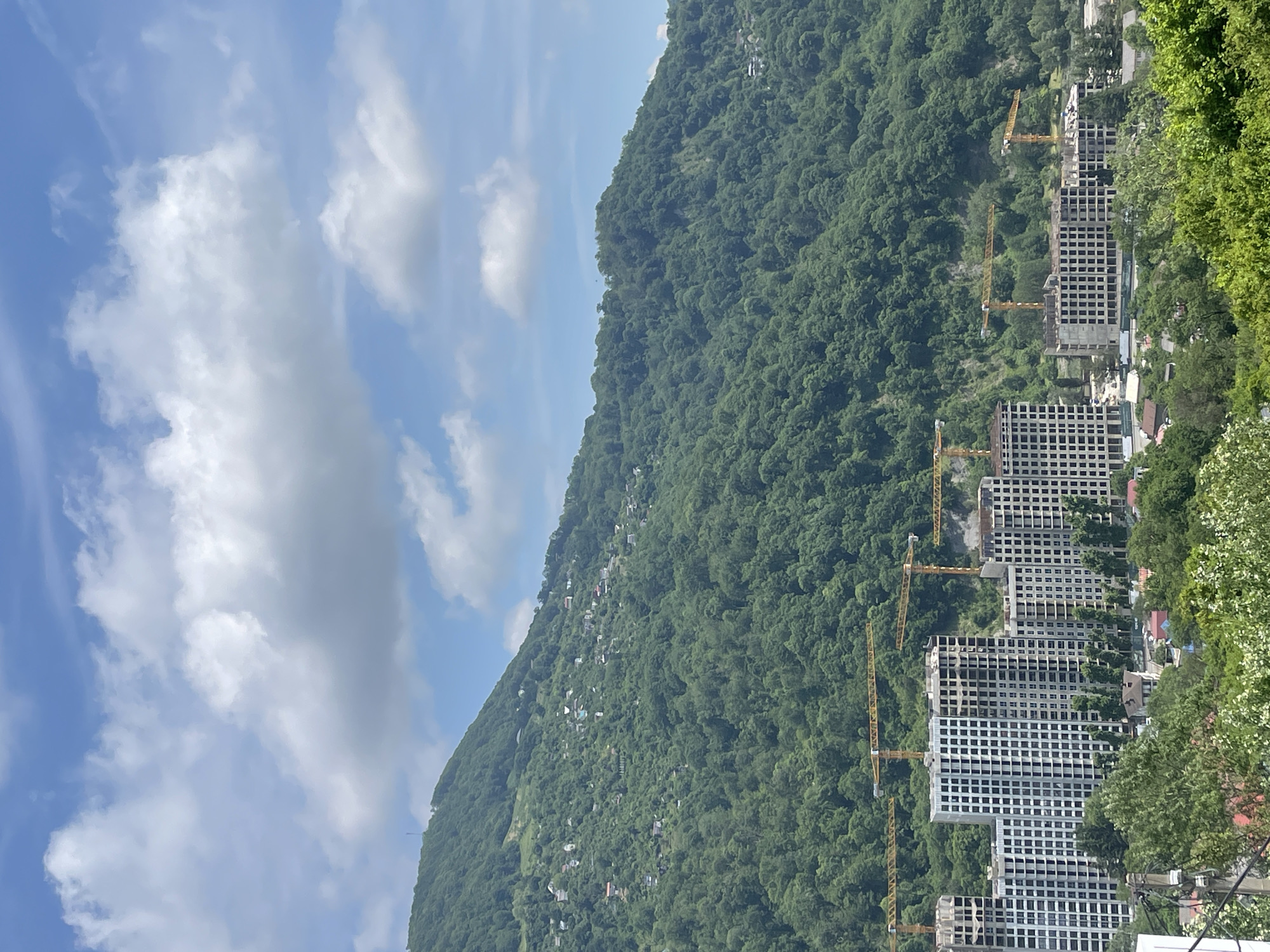 The image displays a panoramic view of a mountainous landscape with significant construction activity in the foreground, identified as the city of Baranovka, Russia.
The upper portion of the scene features a clear blue sky interspersed with scattered white cumulus clouds. Below the sky, a large, densely forested mountain range dominates the background, covered in various shades of green deciduous trees and foliage. Numerous small, light-colored residential structures are visible, sparsely distributed across the lower and mid-slopes of the mountain, particularly on the left side.
In the mid-ground and foreground, a substantial construction project is underway. Multiple multi-story buildings, appearing to be residential complexes, are in various stages of completion. The structures consist primarily of concrete frames, with some buildings having exterior walls and window openings partially finished. At least eight yellow tower cranes are actively operating or positioned across the construction sites, indicating ongoing development. These cranes are positioned above and between the incomplete building structures.
Below the main construction sites, a more established residential area is visible, characterized by smaller buildings with varied roof colors nestled among abundant green trees and shrubs. In the extreme bottom left, elements of utility infrastructure, including power poles and lines, are partially visible. No individuals or specific human activities are discernibly captured in the image.