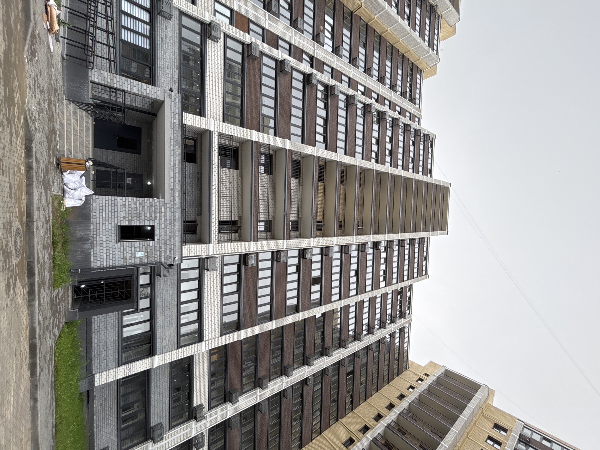 A low-angle, wide shot captures the facade of a modern, multi-story residential building in Blagoveshchensk, Russia. The building features a distinctive architectural style with alternating bands of dark brown brick and lighter colored, possibly concrete or light brick, sections. Numerous windows with dark frames are arranged in rows. Balconies with black railings are visible on some levels. The building appears to be very tall, with its upper floors receding into an overcast sky. The ground level of the building shows an entrance with a dark door, stairs, and a ramp. Several white plastic bags and a wooden trash bin are placed near the entrance. The surrounding area is paved with cobblestones, and patches of green grass are visible along the building's foundation. The scene suggests the building is either new or under construction, given the presence of the bags and the general appearance of the exterior.