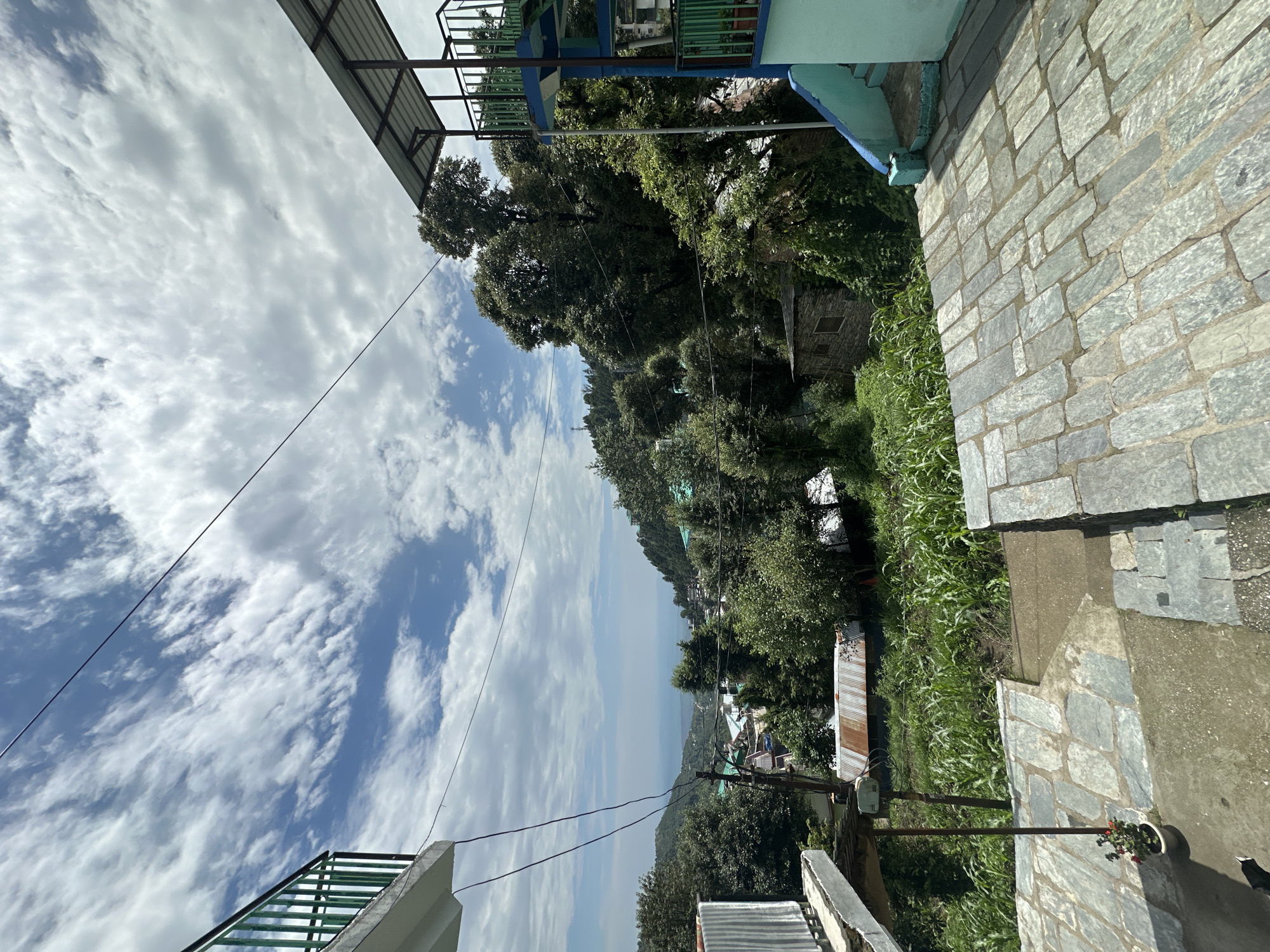 A vertical, eye-level, wide shot captures a scenic view of a town nestled in a valley, framed by lush green trees and a cloudy sky. In the foreground, a paved area with large, flat stone tiles leads to a set of stairs. To the left, a white balcony with a green railing juts out from a building. To the right, a multi-story building with blue and green accents and a metal roof is visible, surrounded by dense foliage. Beyond the immediate structures, the town unfolds with a mix of houses and buildings, interspersed with trees. In the distance, a hazy body of water, possibly the sea or a large lake, is visible, bordered by rolling hills. The sky is filled with a dramatic display of white, fluffy clouds against a bright blue backdrop. The overall scene evokes a sense of peacefulness and natural beauty, with human habitation integrated into the landscape.