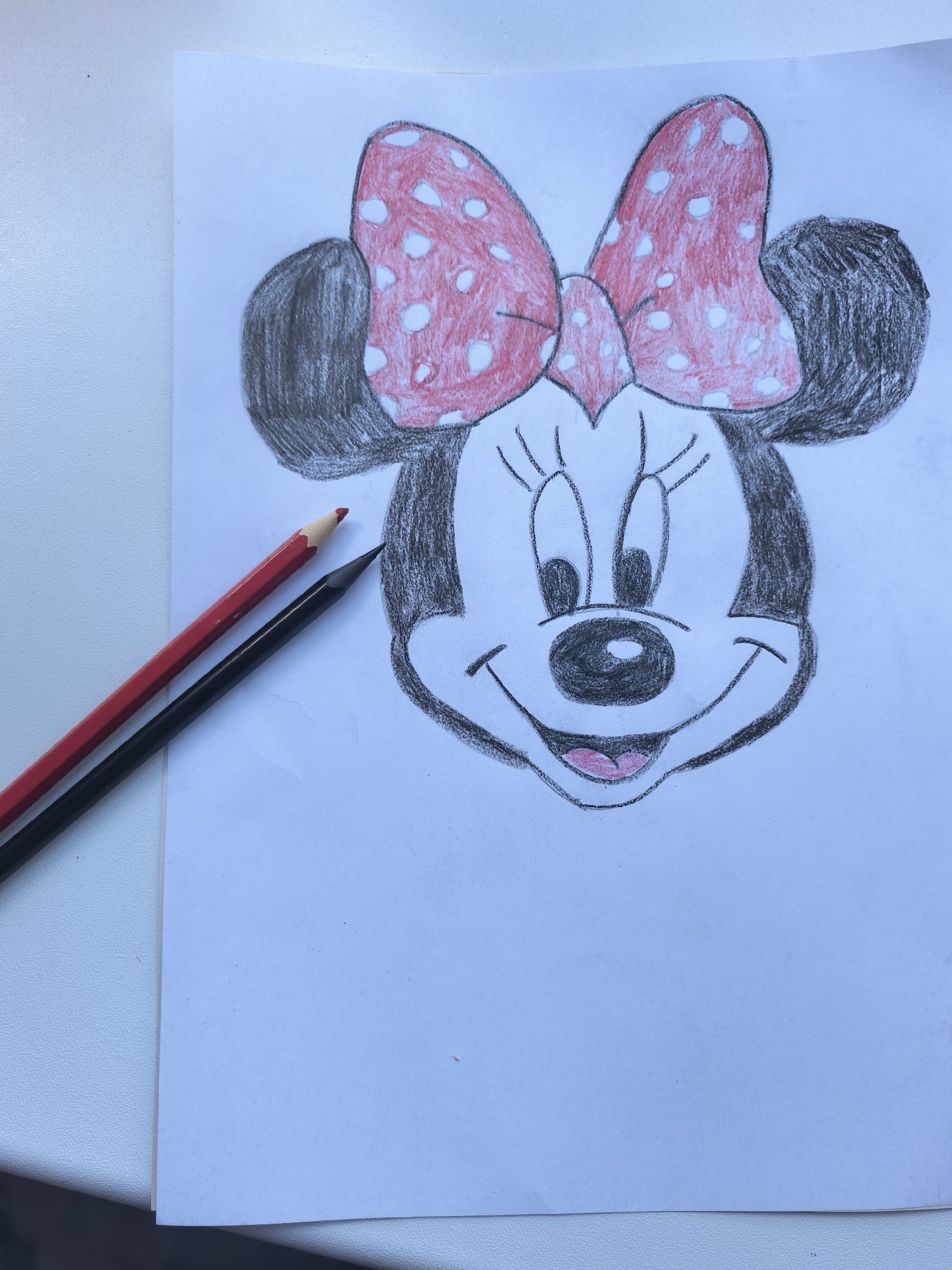 The image shows a drawing of Minnie Mouse's face on a piece of white paper. Minnie Mouse is depicted with her signature red and white polka dot bow. Her ears are black and are drawn with charcoal or graphite. Her face is outlined in black, with black eyes, a black nose, and a pink mouth. The drawing appears to be done with colored pencils, with red used for the bow and pink for the mouth, and black and gray for the ears and facial features. Two colored pencils, one red and one black, are lying on the paper next to the drawing. The drawing is the main subject of the image, and the pencils are secondary objects. The background is a plain white surface, suggesting an indoor setting. There is no visible text in the image. The overall mood of the drawing is cheerful and playful, as conveyed by Minnie Mouse's smiling expression. The image does not provide any clues about the time of day, weather, or specific location within Sterlitamak, Russia, other than that the drawing is being presented on a white surface.