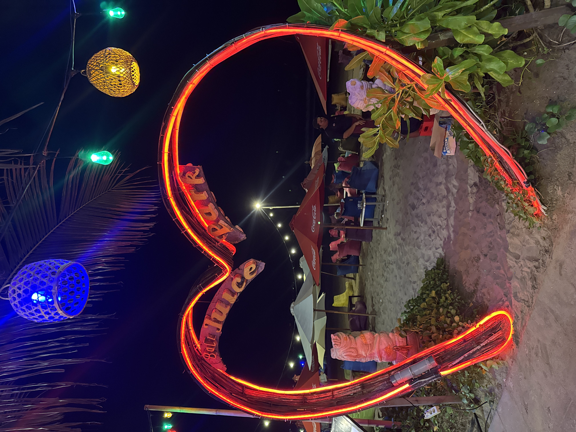 The image depicts a vibrant beachside scene at night in Kuta, Indonesia. Dominating the foreground is a large, illuminated heart-shaped neon sign with red neon tubing. Inside the heart, a wooden sign reads "Bali Juice Park." The sign is positioned on a sandy beach, with lush green foliage growing around its base.
Behind the heart, the beach is dotted with various seating arrangements, including beanbag chairs in an array of colors (pink, blue, green, yellow) and beach umbrellas, some of which are red and bear the Coca-Cola logo. There are also a few white and light-colored umbrellas. A pale pink, statue-like object, possibly a carved figure, stands near the left side of the heart.
In the background, the dark night sky is visible, punctuated by strings of colorful lights. One prominent light is a blue woven lantern hanging from a palm frond. Other lights include green, pointed bulbs and a yellow woven spherical lantern. A few people are faintly visible in the background, one standing near the seating area. The overall atmosphere is festive and inviting, characteristic of a beach bar or entertainment venue at night.