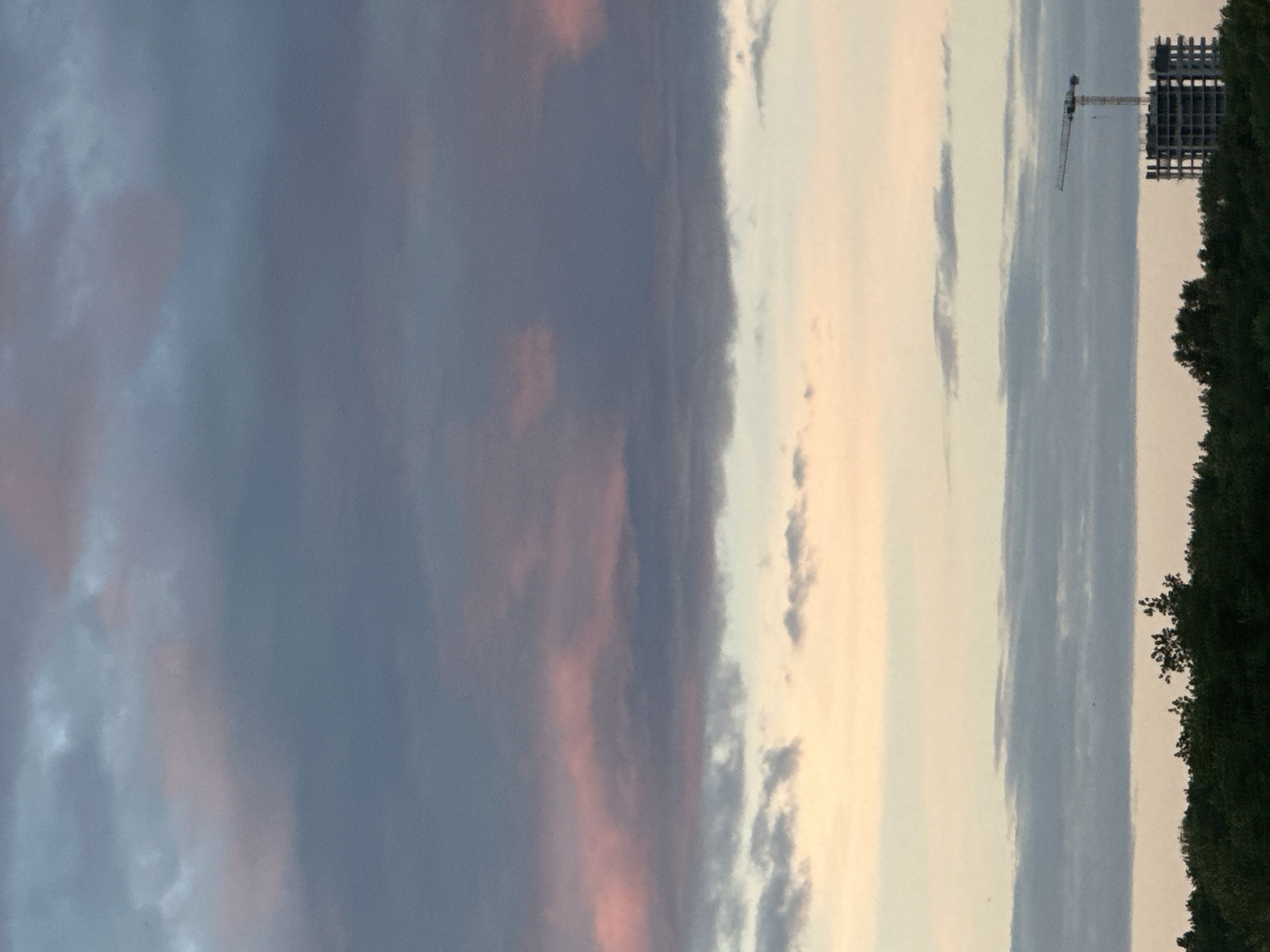 The media file depicts a view of the sky with clouds, a construction crane, and a partially constructed building. The sky exhibits hues of grey, pink, and yellow, suggesting a time around sunrise or sunset. The clouds are scattered, with some appearing darker and more dense than others. On the right side of the frame, there is a silhouette of trees, forming a dark line against the lighter sky. In the distance, a large construction crane is visible, along with a building under construction, characterized by its skeletal structure. The scene appears to be set in an outdoor environment, possibly on the outskirts of a city given the presence of construction. There is no discernible text in the image. The overall mood is calm, with the colors of the sky creating a serene atmosphere.