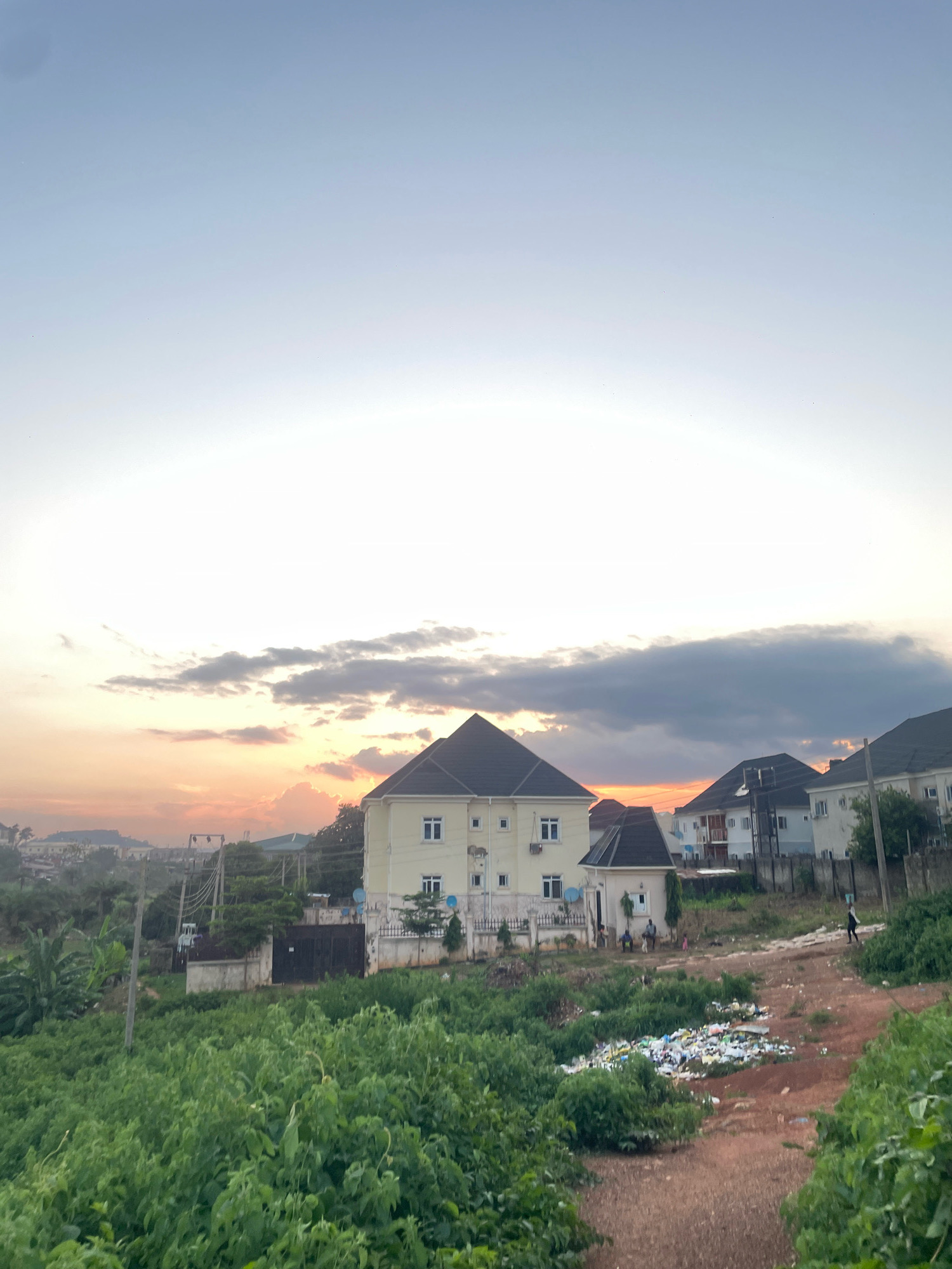 The image depicts a residential area on the outskirts of Abuja, Nigeria, likely during either sunrise or sunset, given the warm hues in the sky and the soft lighting. In the foreground, dense green vegetation dominates the left side of the frame, with a dirt path winding through it on the right. This path leads to a pile of accumulated refuse, including plastic bottles and other debris, situated on a patch of disturbed earth.
Further back, a cluster of modern residential buildings is visible. The most prominent is a multi-story, pale yellow house with a dark pitched roof, featuring multiple windows and balconies. To its right and slightly behind, other houses of similar architectural style, characterized by light-colored facades and dark roofs, are present. Some of these buildings show satellite dishes on their roofs.
In the mid-ground, utility poles with visible wiring are present, suggesting the presence of electricity infrastructure. There are also gates and low walls defining property boundaries. A few individuals are discernible in the scene. Near the pale yellow house, two men are seated, and a small child is standing nearby, while a woman with an object on her head walks along the dirt path in the mid-right of the image. The overall impression is a developing urban environment where residential construction coexists with natural landscape and issues of waste management. The distant background shows a hazy, hilly landscape.