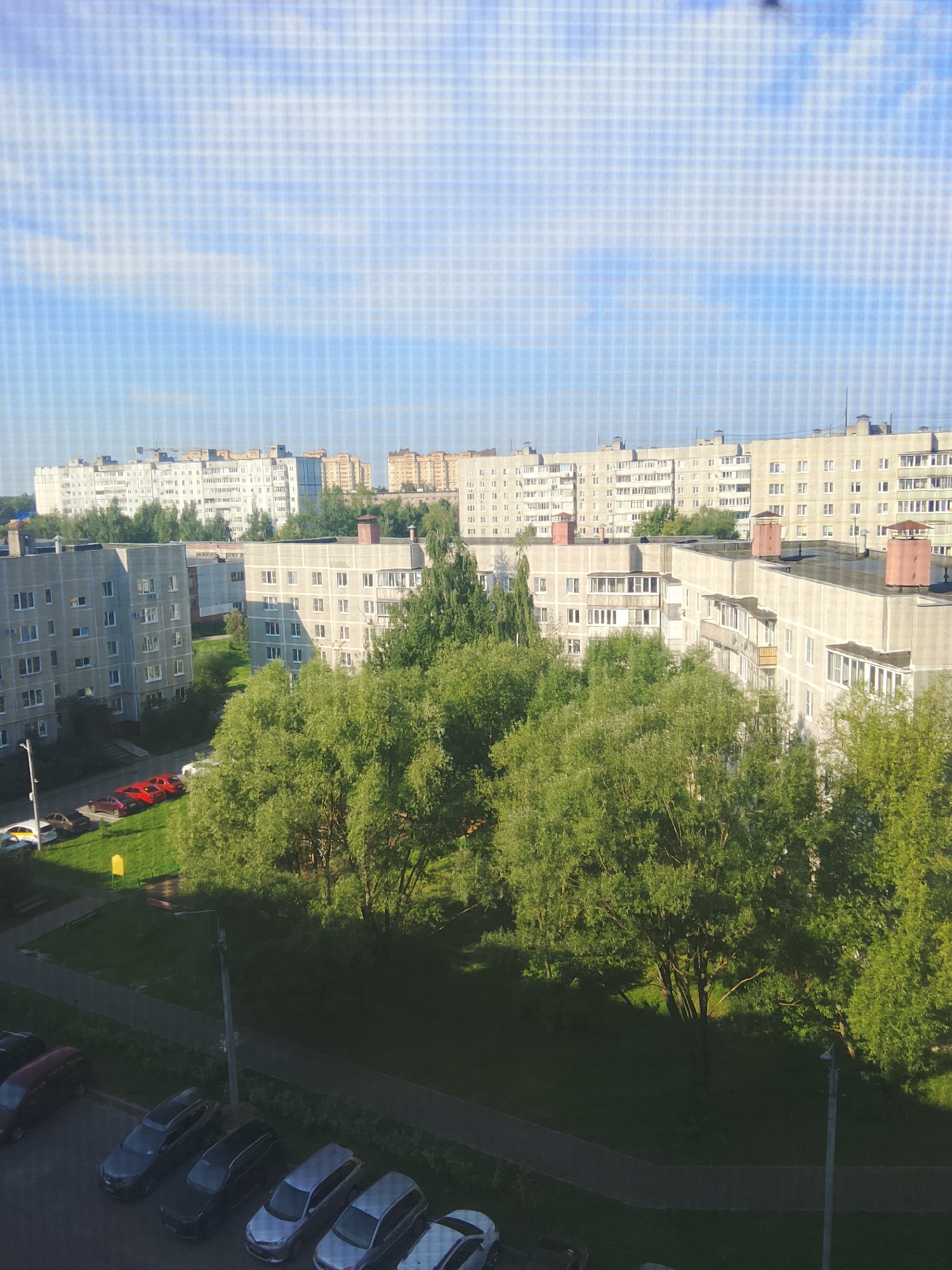 An aerial view of a residential area in Yegoryevsk, Russia, features a cluster of multi-story apartment buildings interspersed with lush green trees. The buildings are primarily light-colored, with visible windows and balconies. In the foreground, several cars are parked on asphalt surfaces adjacent to green lawns. One section shows a row of cars, including dark-colored sedans and silver hatchback-style vehicles. Further back, a few red cars are parked on a green, grassy area. The sky above is a light blue with scattered clouds. The scene appears to be taken on a bright, sunny day, with shadows cast by the buildings and trees. The image captures a typical urban residential environment.