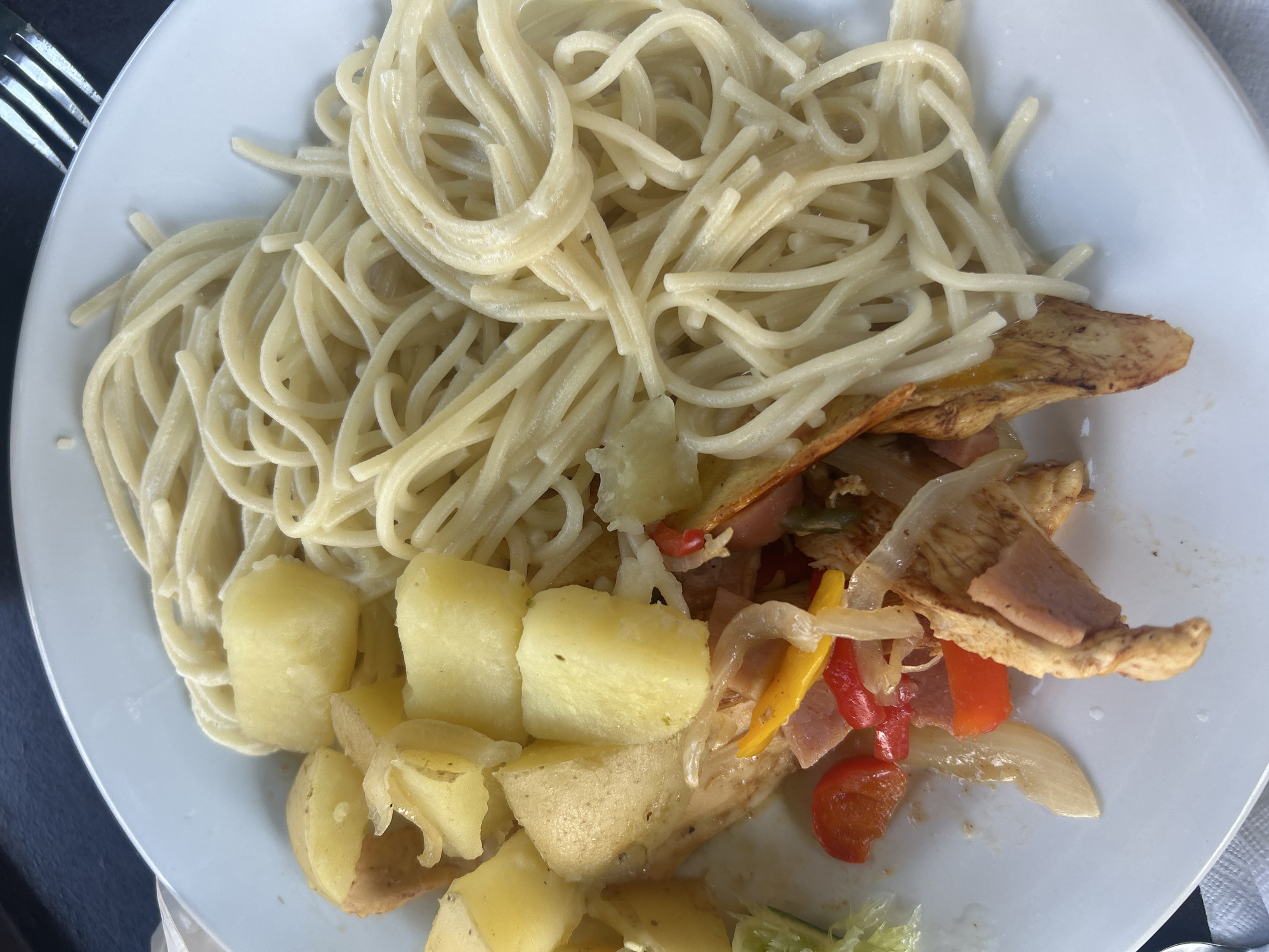 A top-down view of a white plate filled with spaghetti, potatoes, and stir-fried vegetables, possibly chicken or another light-colored meat. The spaghetti takes up the top left portion of the plate, while the potatoes occupy the bottom left. The stir-fried vegetables are on the right and contain red and yellow peppers, onions, ham, and possibly chicken. The plate is placed on what appears to be a dark surface. A fork is partially visible on the top left. The photo was potentially taken in San Bernardino Tlaxcalancingo, Mexico.