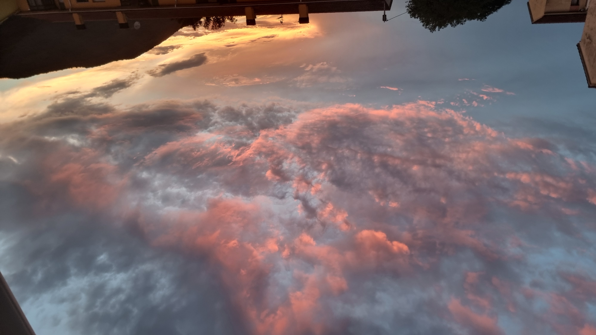 The image shows a sunset with colorful clouds over the town of Sant Joan Les Fonts, Spain. The sky is filled with dramatic clouds displaying hues of pink, orange, and gray. The lower portion of the image features the rooftops of buildings, a dark silhouetted tree, and the outline of a hill. The scene captures a serene evening atmosphere.
