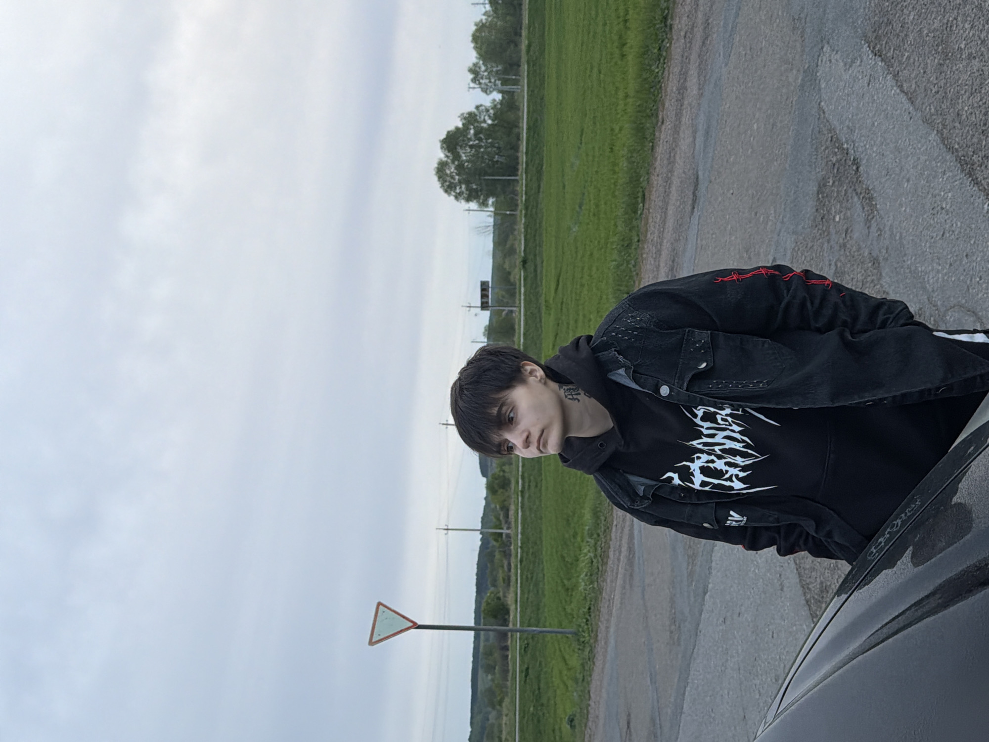 A medium shot depicts a person with short dark hair wearing a black hoodie with a white graphic design and a denim jacket, standing next to a car. They are positioned on a paved road adjacent to a grassy field. In the background, utility poles are visible along with dense foliage. A road sign is also present. The sky is overcast. Based on available data, the location is Azyakul', Russia.