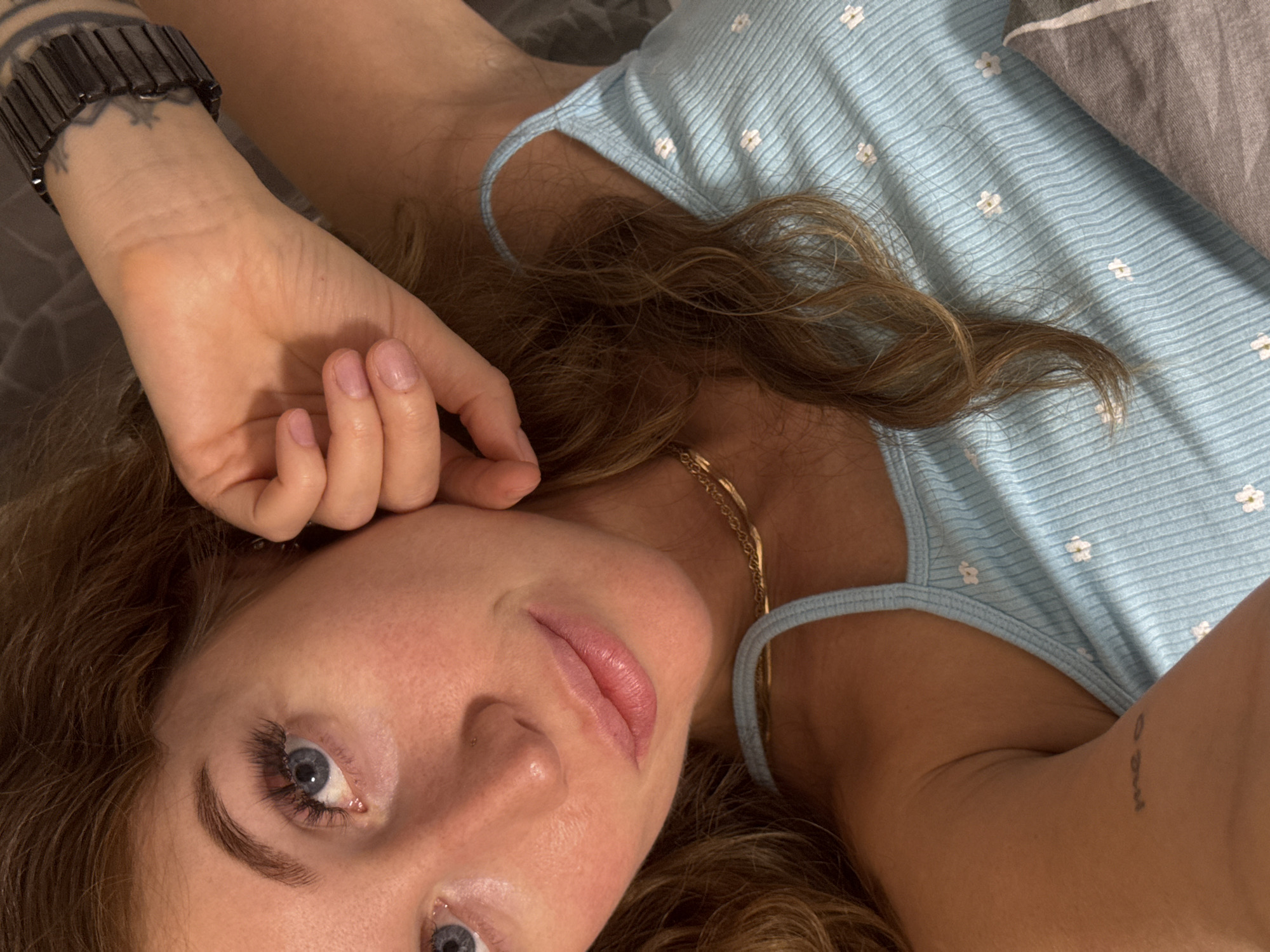 A woman with light brown hair and blue eyes is lying on her back, wearing a light blue tank top with white flower patterns and a gold chain necklace. Her right hand is raised near her face, showcasing manicured fingernails and a black watch with a tattoo on her wrist. A small tattoo is visible on her upper left arm. The background consists of bedding with a light blue and gray pattern. There is no indication of this being in the city of Chaikovsky, Russia.