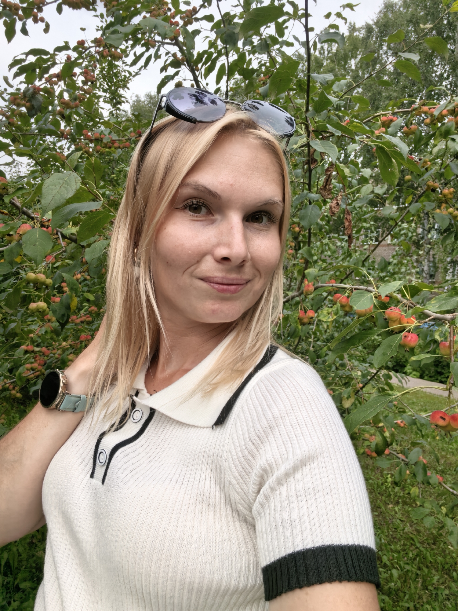 A young Caucasian woman with blonde hair and sunglasses resting on her head is captured in a medium selfie. She is smiling and looking at the camera, positioned in front of a tree laden with small red and green fruits. The woman is wearing a white ribbed polo shirt with black trim and a smartwatch with a light blue band on her left wrist. The background consists of lush green foliage and the overcast sky, suggesting an outdoor setting likely in Sterlitamak, Russia.