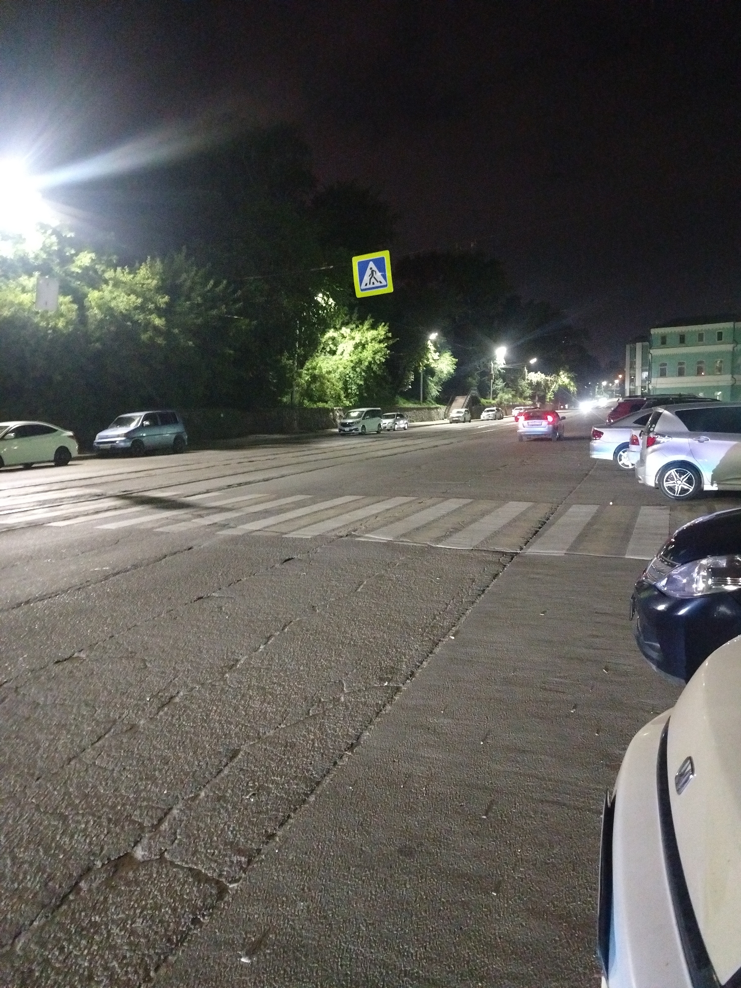 An exterior night-time shot captures a city street in Irkutsk, Russia, with a pedestrian crossing illuminated by streetlights and the headlights of parked and moving vehicles. The road surface exhibits cracks and is flanked by trees and a building. Several cars are parked along the roadside and in the street, with some in motion. A pedestrian crossing sign, depicting a walking figure, is visible. The lighting conditions create strong contrasts and shadows.