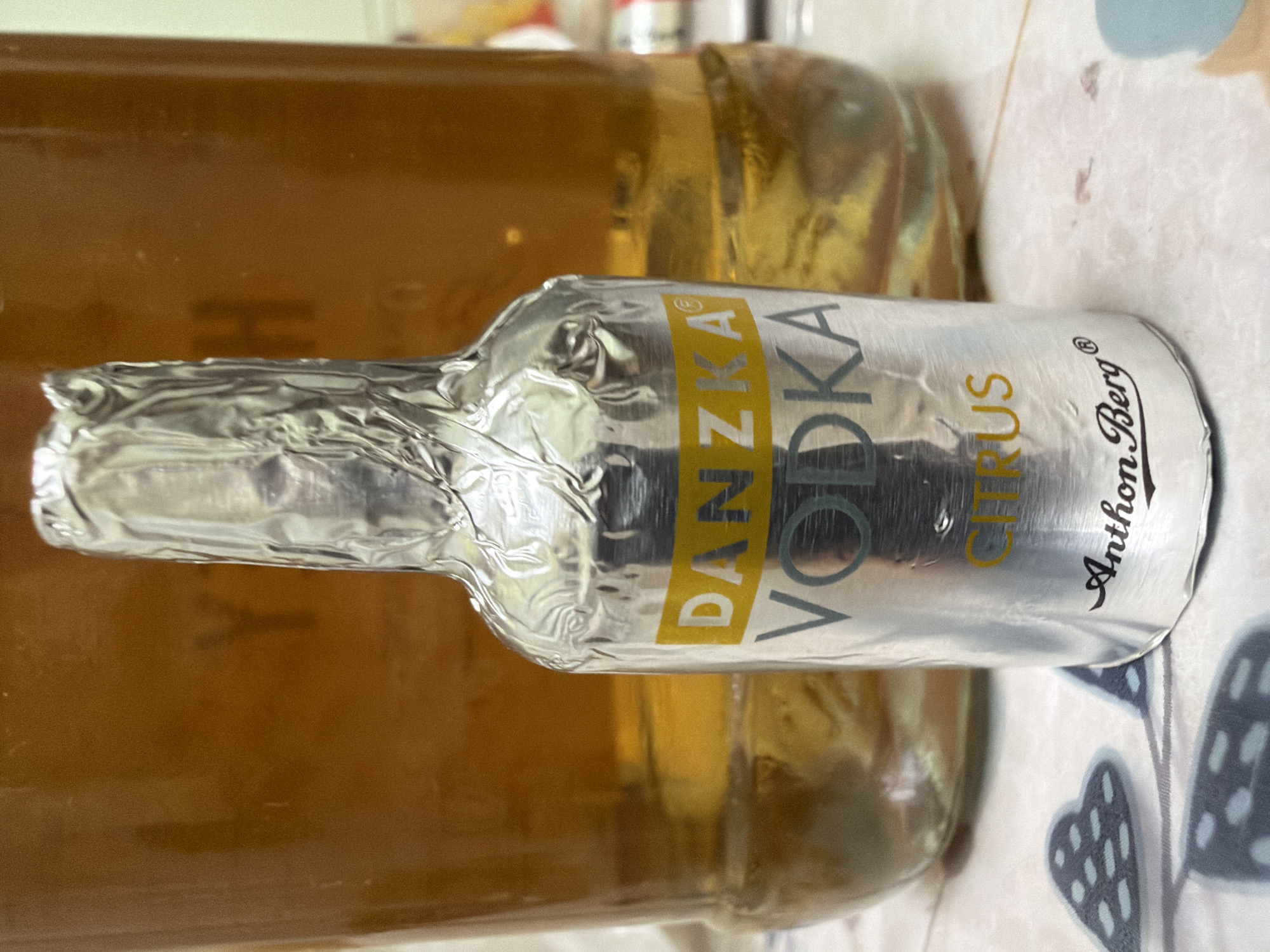 A close-up, eye-level, indoor shot shows a miniature bottle of Danzka Vodka Citrus wrapped in silver foil. The bottle stands in front of a larger glass bottle filled with a golden-brown liquid, possibly another alcoholic beverage or tea. The background bottle appears to have dark, indistinct lettering visible through the liquid. The foreground bottle has a yellow band with the brand name "DANZKA" printed in black. Below that, "VODKA" is printed in large, grey lettering. At the bottom, "CITRUS" is written in yellow, and the signature "Anthon Berg" is in black cursive. The scene is set on a patterned surface, possibly a tablecloth, with a blue and grey heart-shaped design. The lighting is somewhat dim, creating reflections on the foil-wrapped bottle. The image provides no information about its location within Kyiv, Ukraine.
