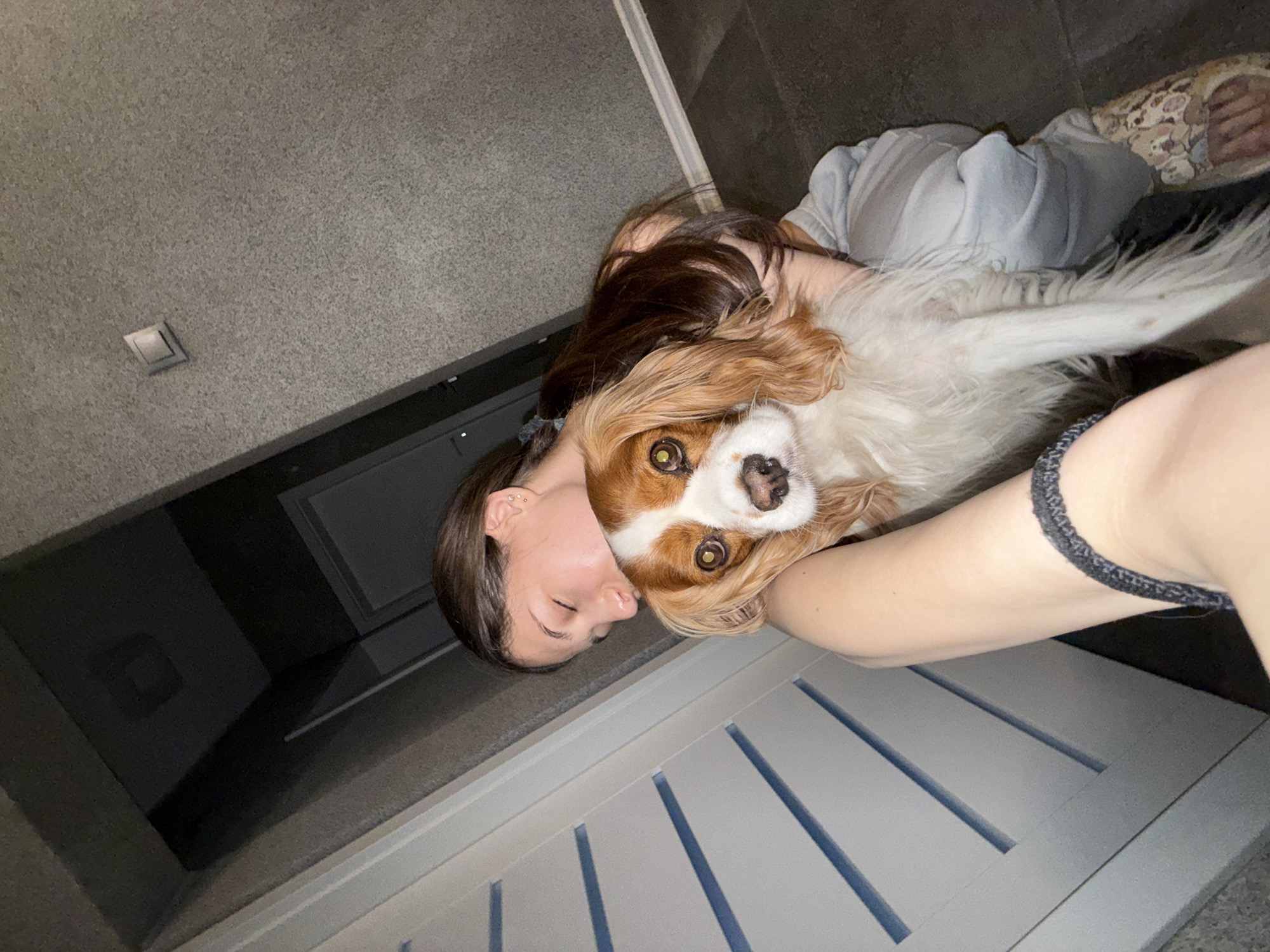 A woman with long brown hair is lying down with a Cavalier King Charles Spaniel dog. She is holding the dog closely, with her face near the dog's head. The dog is looking directly at the camera with its large brown eyes. The woman is wearing a grey tank top and light-colored pajamas. A portion of her feet is visible, wearing patterned slippers. They appear to be lying on or near a grey-colored wooden surface. The background consists of a grey wall and a dark area, possibly a piece of furniture or a wall. The overall lighting is dim. The specific location in Sofia, Bulgaria cannot be determined from this image.
