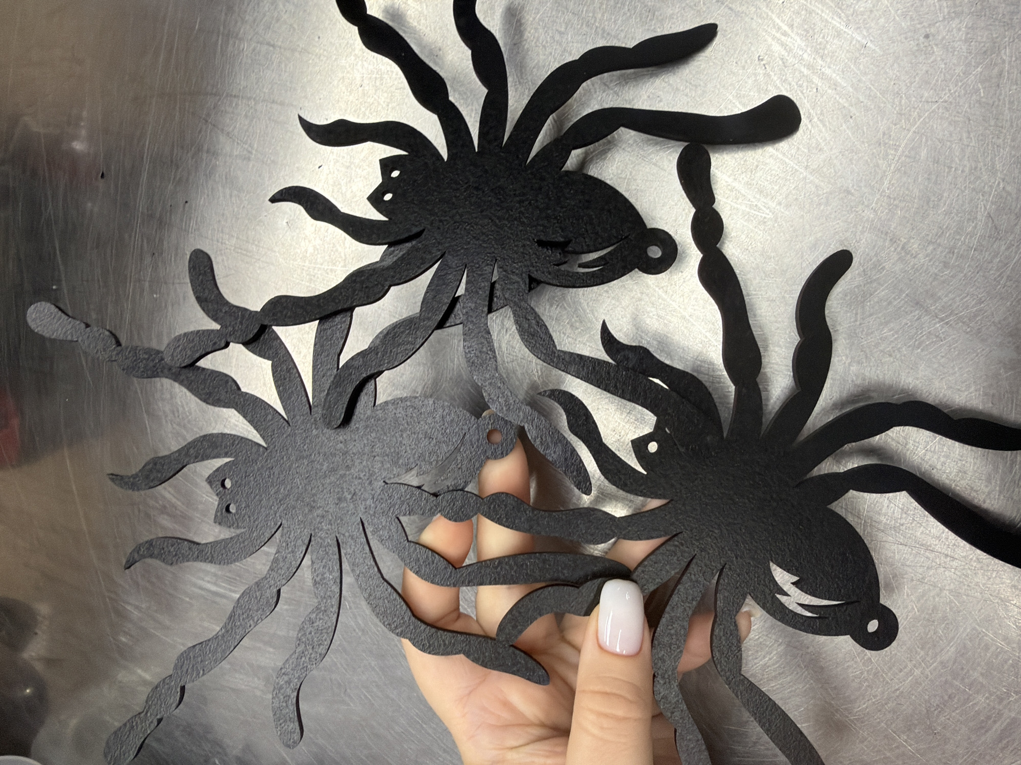 Three black paper spider decorations are displayed on a brushed metal surface. A human hand with white nail polish is holding the leftmost spider. The spiders have eight legs and two circular eye cutouts. One spider has a jagged lightning bolt shape cut out of its abdomen. The surface appears to be a countertop or table with visible scratches. The location is unknown.