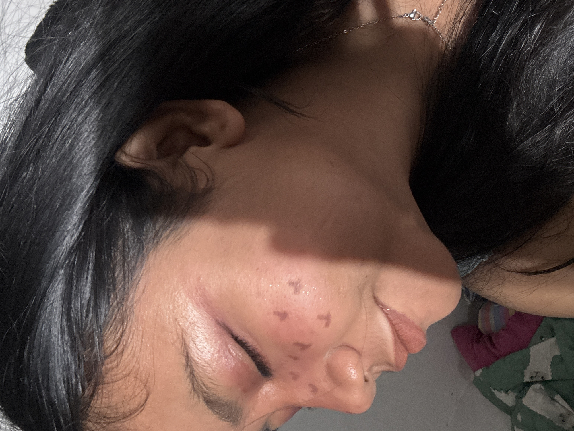 A close-up profile view of a young woman's face with dark hair, taken indoors, possibly in North Jakarta, Indonesia. She is looking down and to the left with her eyes closed. Her makeup includes pink eyeshadow and what appear to be small, dark heart-shaped markings on her cheeks and nose. A silver necklace is visible around her neck. In the background, a white wall and a portion of a patterned blanket are visible. The lighting suggests an artificial indoor light source.