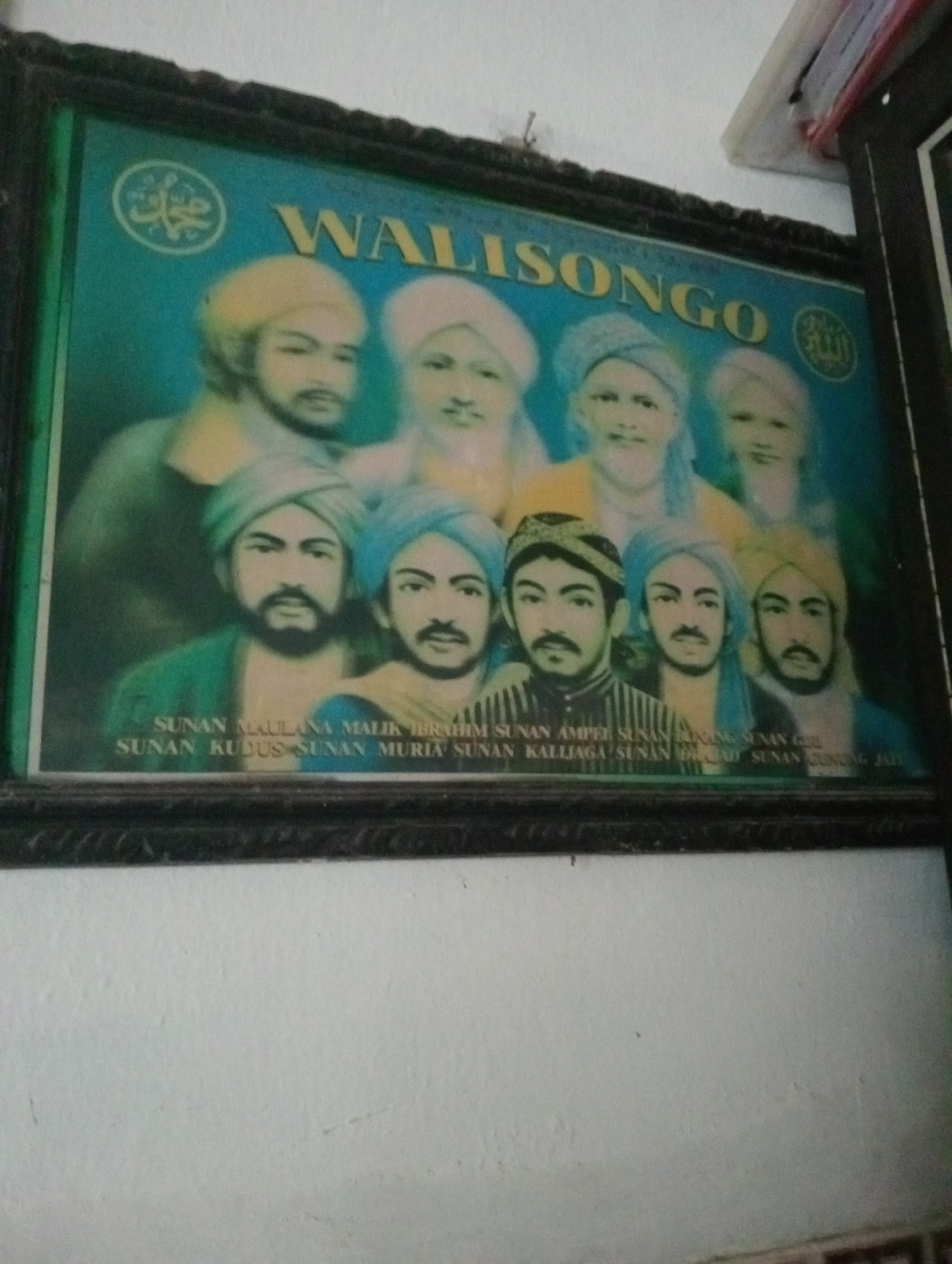 A framed portrait depicting the Wali Songo, a group of nine revered Islamic saints in Indonesia, is prominently displayed on a white wall. The portrait features an assembly of men, each with distinct facial features and head coverings, against a teal background. The text "WALISONGO" is emblazoned across the top of the portrait. Below the figures, a list of names, likely identifying each saint, is partially visible. The overall image is a reproduction, likely a print or poster, housed within a dark frame with ornate detailing. The setting appears to be an interior space, possibly a home or public building in Desa Silo, Indonesia, given the presence of the portrait which is culturally significant in the region.
