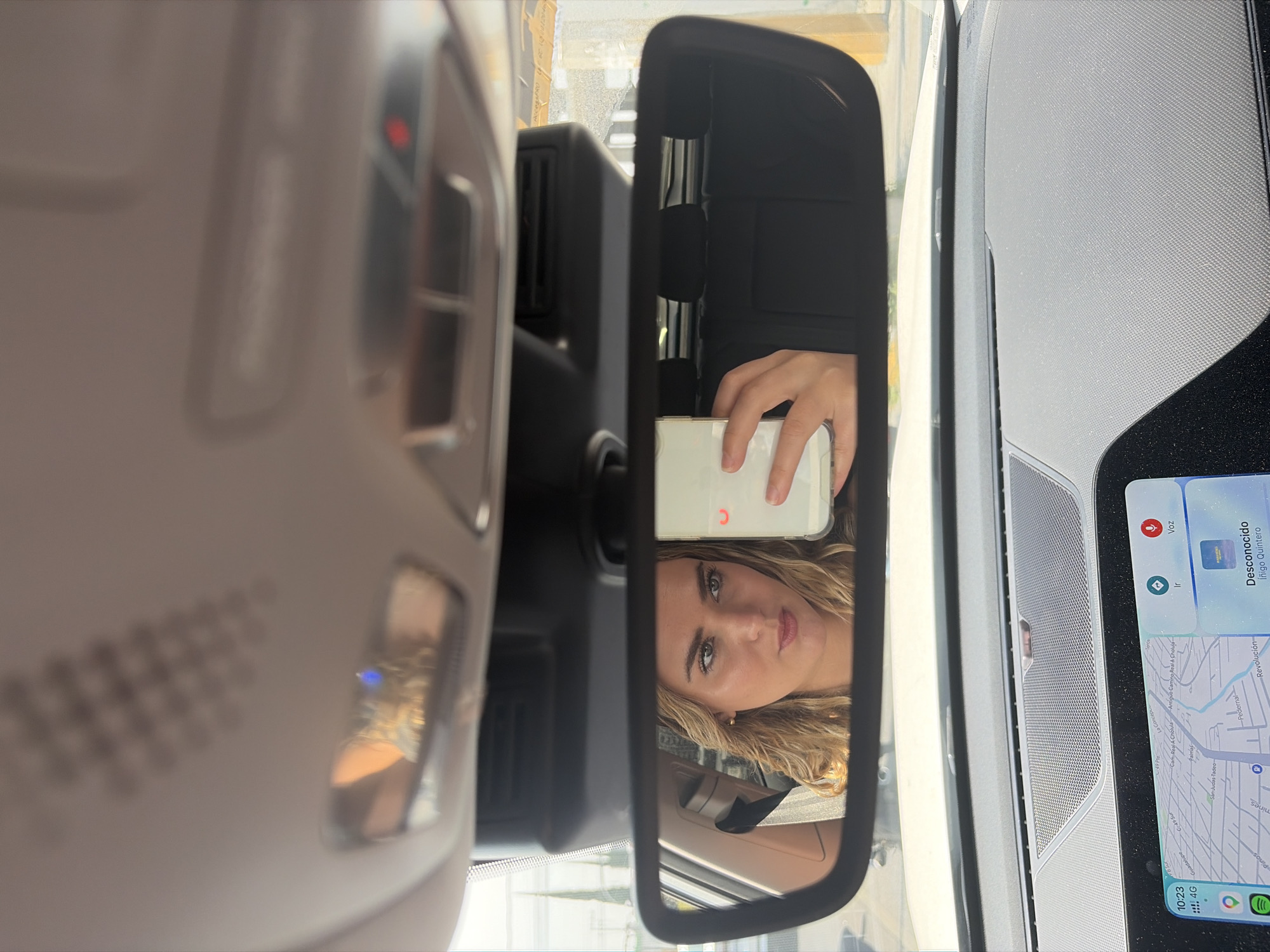 The image captures a reflection in a car's rearview mirror, showing a woman with blonde hair looking directly forward, possibly at her smartphone. She holds a white smartphone with a clear case in her right hand, positioned in front of her face, suggesting she might be taking a selfie or checking her phone.
The scene is set inside a car during the daytime, indicated by the bright light entering through the windshield. To the left of the mirror, the car's overhead console is visible, featuring buttons and a small illuminated red indicator light. To the right, part of the car's dashboard with a textured speaker grille and an infotainment screen is seen.
The infotainment screen displays a map with several street names, including "Antiguo Camino Real A Cholula," "Revolución," "Pedernal," "Palmas," "C-49 Sur," "Guadalupe," and "San Judas Tadeo." These street names, along with the Spanish text such as "Voz," "Ir," and "Desconocido," confirm the location to be Puebla, Mexico, specifically around the Cholula area. The screen also shows the time "10:23," "4G" connectivity, a Spotify icon, and a contact listed as "Desconocido Iñigo Quintero." The woman appears to be in the driver's seat.