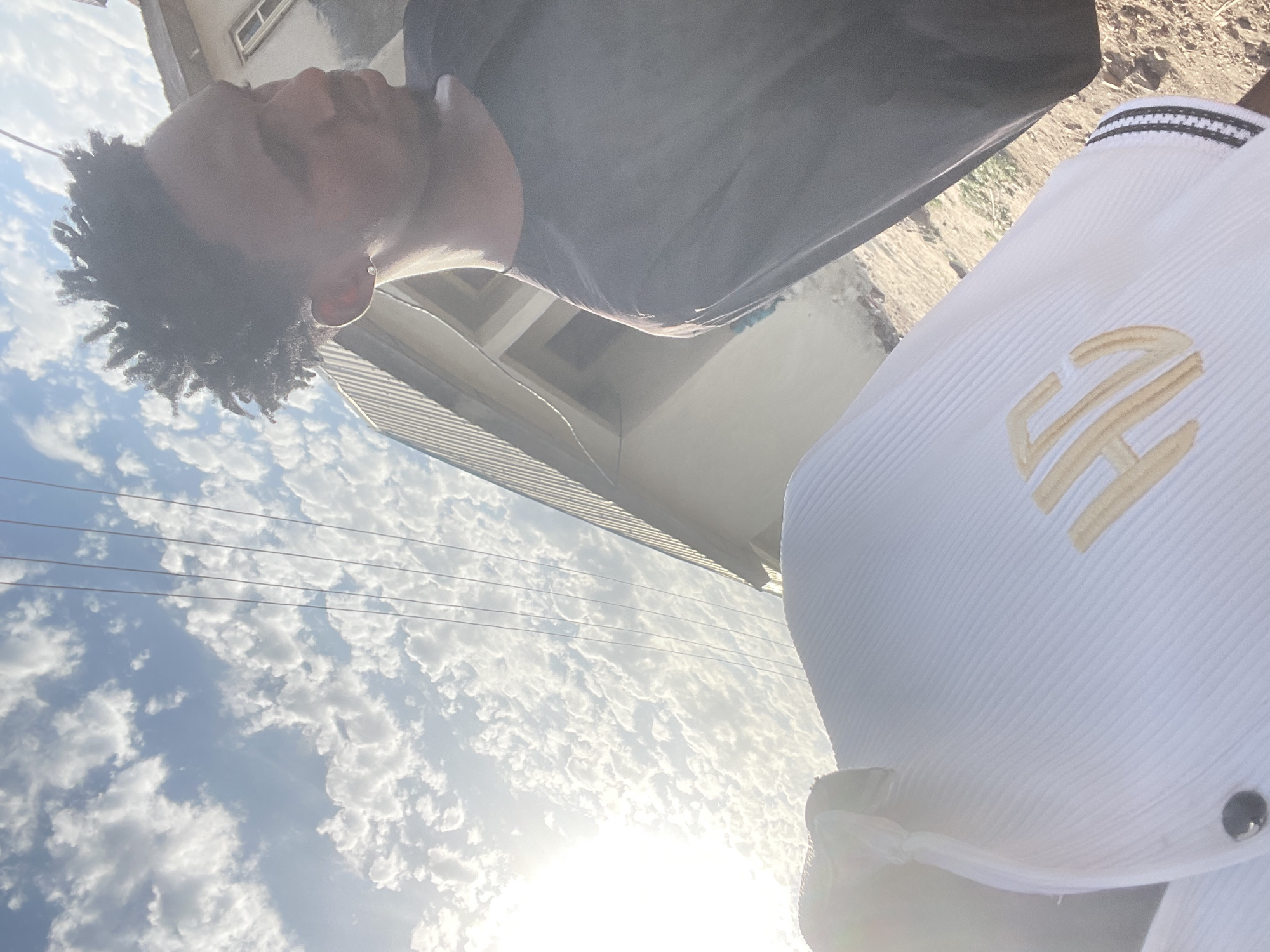 A low-angle shot shows a dark-skinned person with dreadlocks wearing a black shirt. Next to them is a person wearing a white jacket or shirt with gold letters, possibly initials, on it. The sky is visible, with a dense cloud cover and a bright sun shining through. Power lines cut across the sky. Parts of buildings are visible in the background. It is impossible to verify if the location is Lokoja, Nigeria from this image alone.