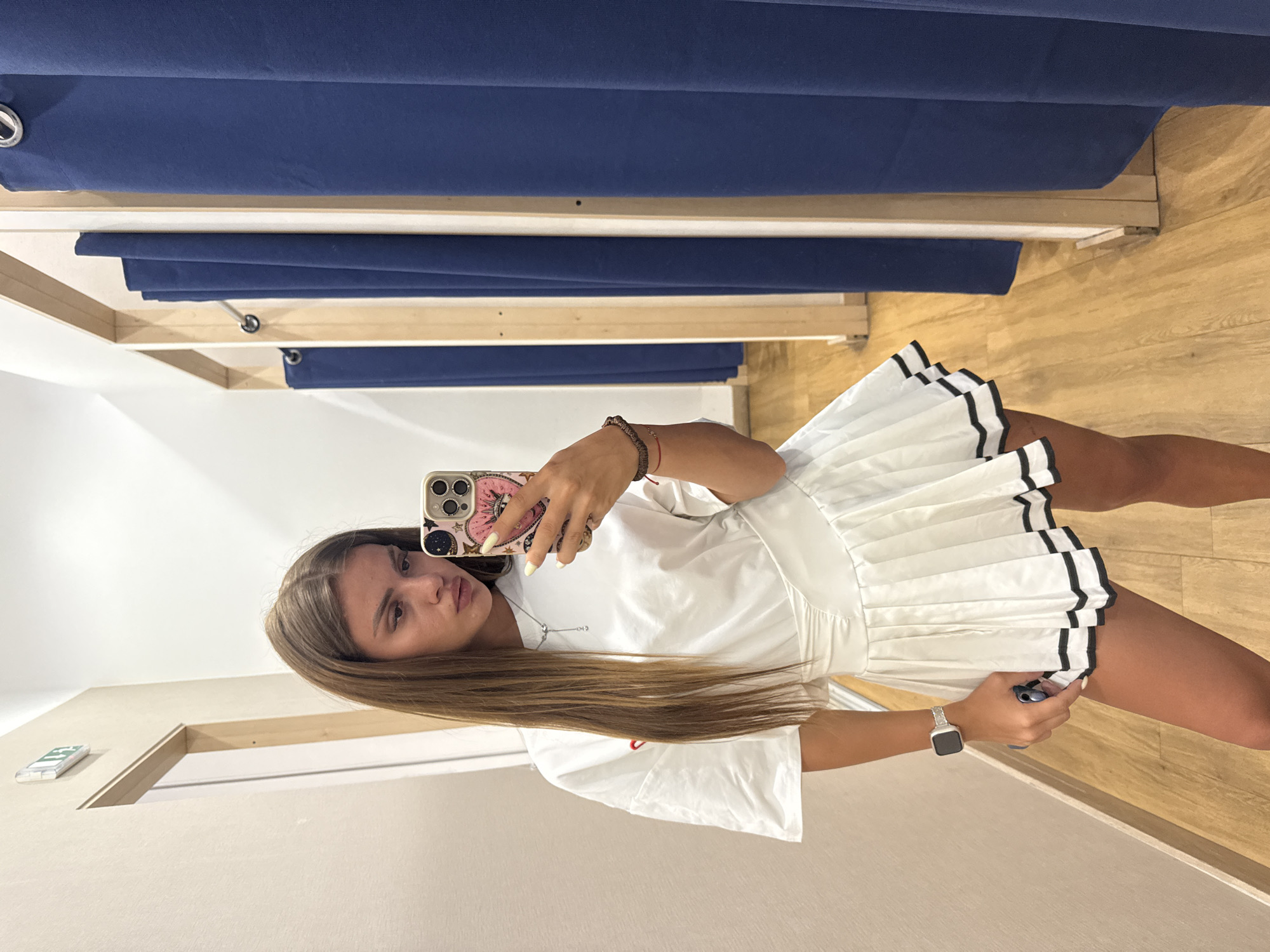 A young woman is taking a selfie in a fitting room. She is wearing a white t-shirt dress with a pleated skirt. The skirt has black trim at the hem. She has long, brown hair and is wearing a silver watch on her left wrist. She is holding a smartphone in her right hand, which has a pink case with astrological symbols on it. A red string bracelet is visible on her left wrist. The fitting room has blue curtains and wooden floors. There is a mirror reflecting the scene. The overall impression is casual and youthful. The lighting suggests it is daytime. There is no visible text in the image.