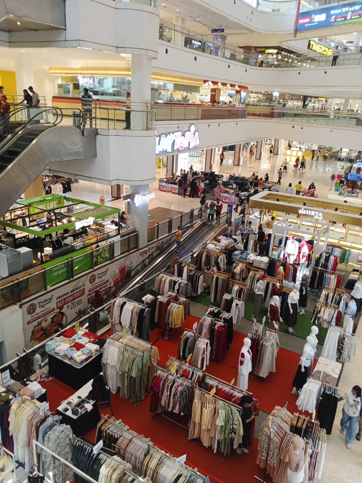 This is a high-angle, medium shot of the interior of a multi-level shopping mall, likely in the City of Medan, Indonesia, featuring numerous retail displays and shoppers. In the foreground, several clothing racks are arranged on a red carpeted floor, showcasing a variety of apparel, including dresses, shirts, and skirts. Mannequins dressed in white and red outfits are also visible among the clothing displays.
Further back, escalators are seen connecting different levels of the mall, with people using them. Several food and beverage stalls are present, identified by signs like "LOVE LIFE" and "BOOST." One stall has green awnings and displays food items. Other shops with various signage are visible on higher floors, including what appears to be a "TIMEZONE" gaming arcade and "Lenovo" electronics store.
Shoppers, including families and individuals, are scattered throughout the mall, browsing merchandise, walking, and conversing. The overall environment is bright, with overhead lighting illuminating the spacious interior, and large windows or openings offer glimpses of the mall's upper levels and architecture. The presence of diverse retail outlets and the density of shoppers suggest a busy commercial area.