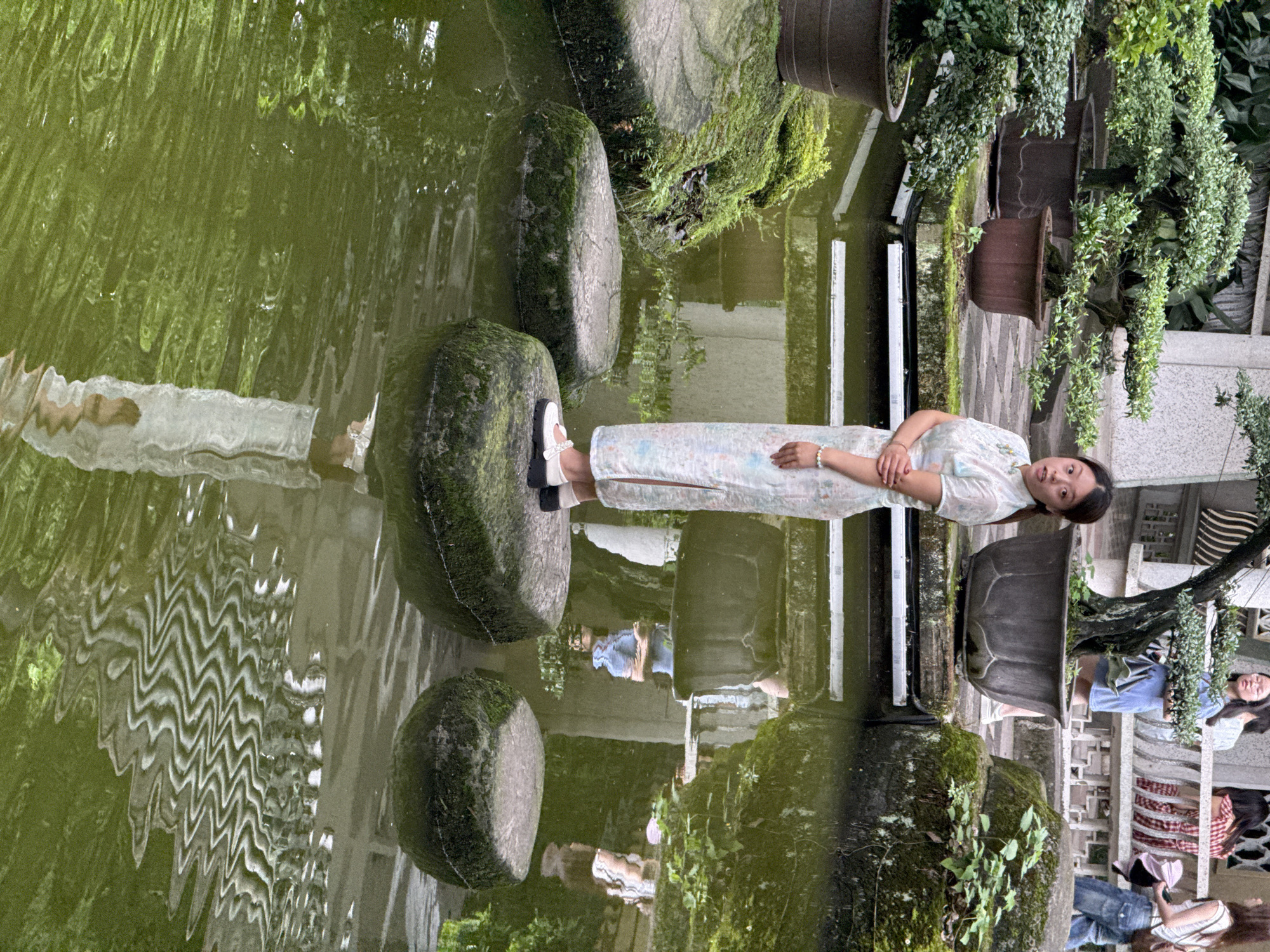 A horizontal eye-level shot captures a woman in Yuzhong District, China, standing on moss-covered stones in a pond. The pond's water is murky green, reflecting the sky and surrounding architecture.
The woman, the focal point of the image, is wearing a light-colored, patterned dress and white shoes with black accents. She is standing with her arms crossed.
Behind the pond, potted plants with cascading greenery are arranged along a ledge. In the background, other people are visible on a tiled platform, standing and sitting, and the architecture is identifiable as traditional Chinese.