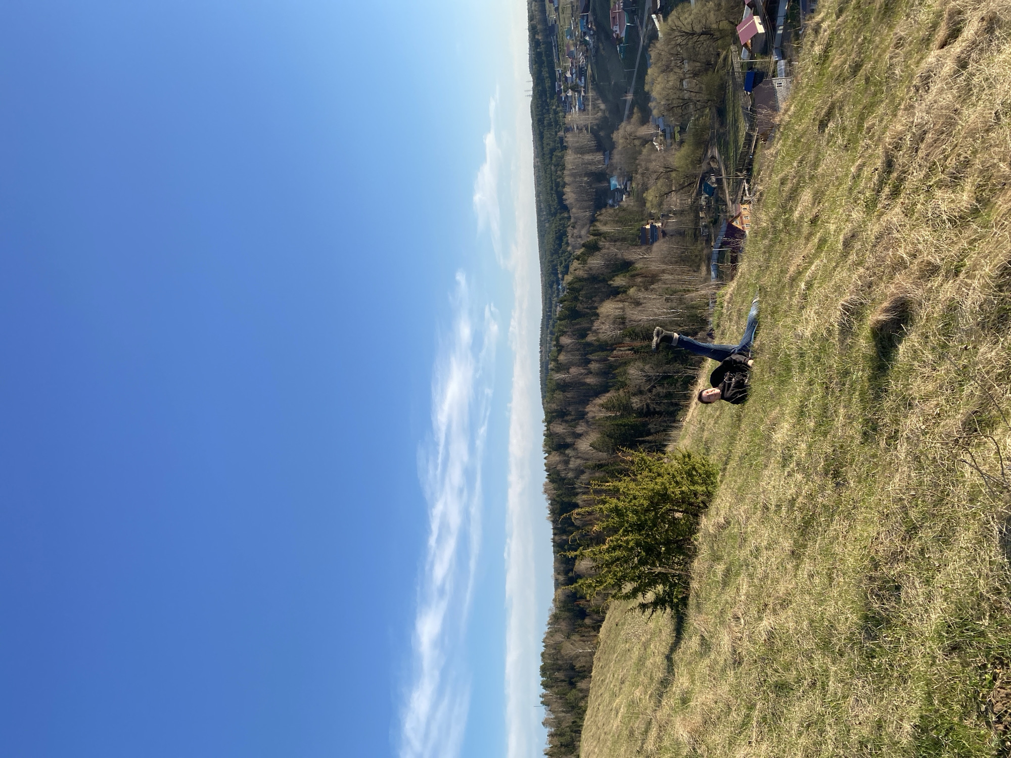 A person is lying on a grassy hillside. The person is wearing jeans, dark shoes, and a dark jacket, and has a dark backpack. The hillside has dry grass and some green bushes. In the background, there is a forest and some buildings, likely part of the city of Ольховка, Russia. Above the forest and buildings is a blue sky with streaks of white clouds.